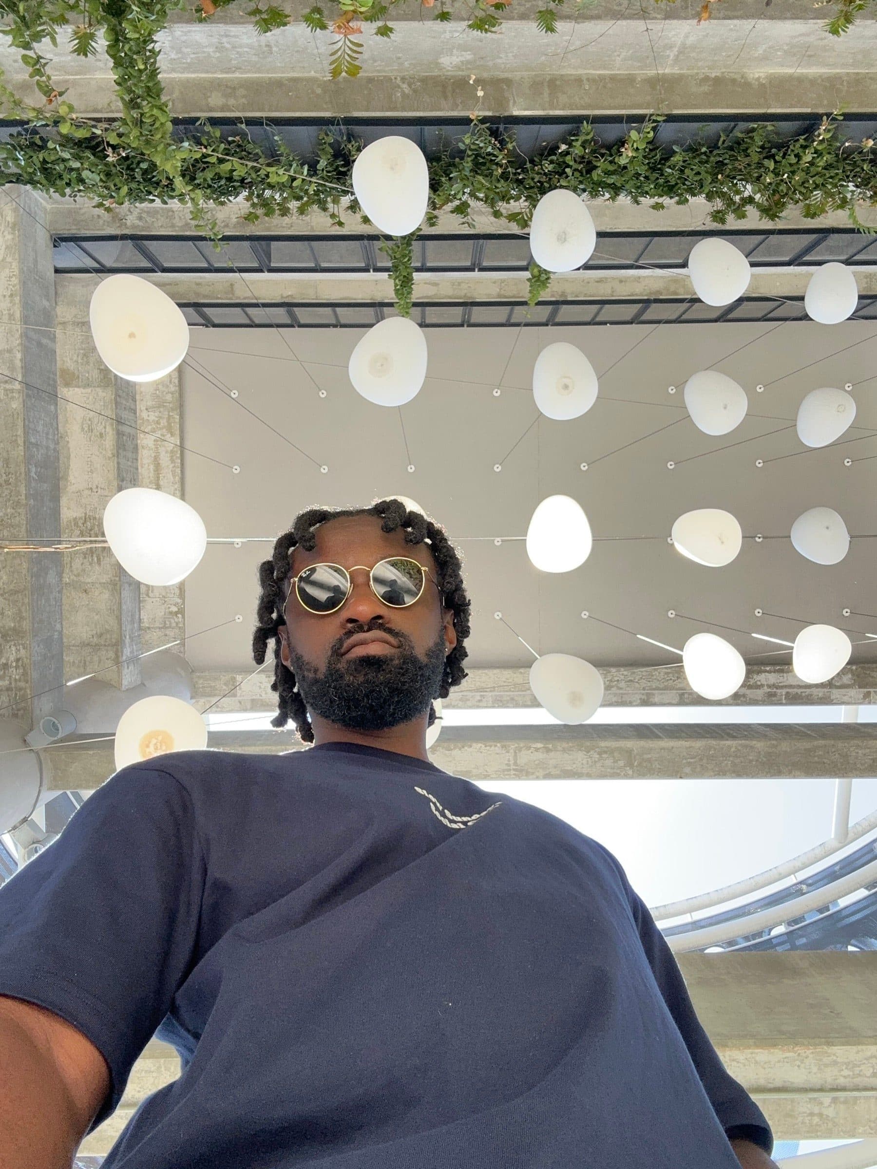 Low-angle portrait of Derek Adu in round sunglasses and a navy t-shirt beneath a ceiling of white pendant lights and hanging greenery