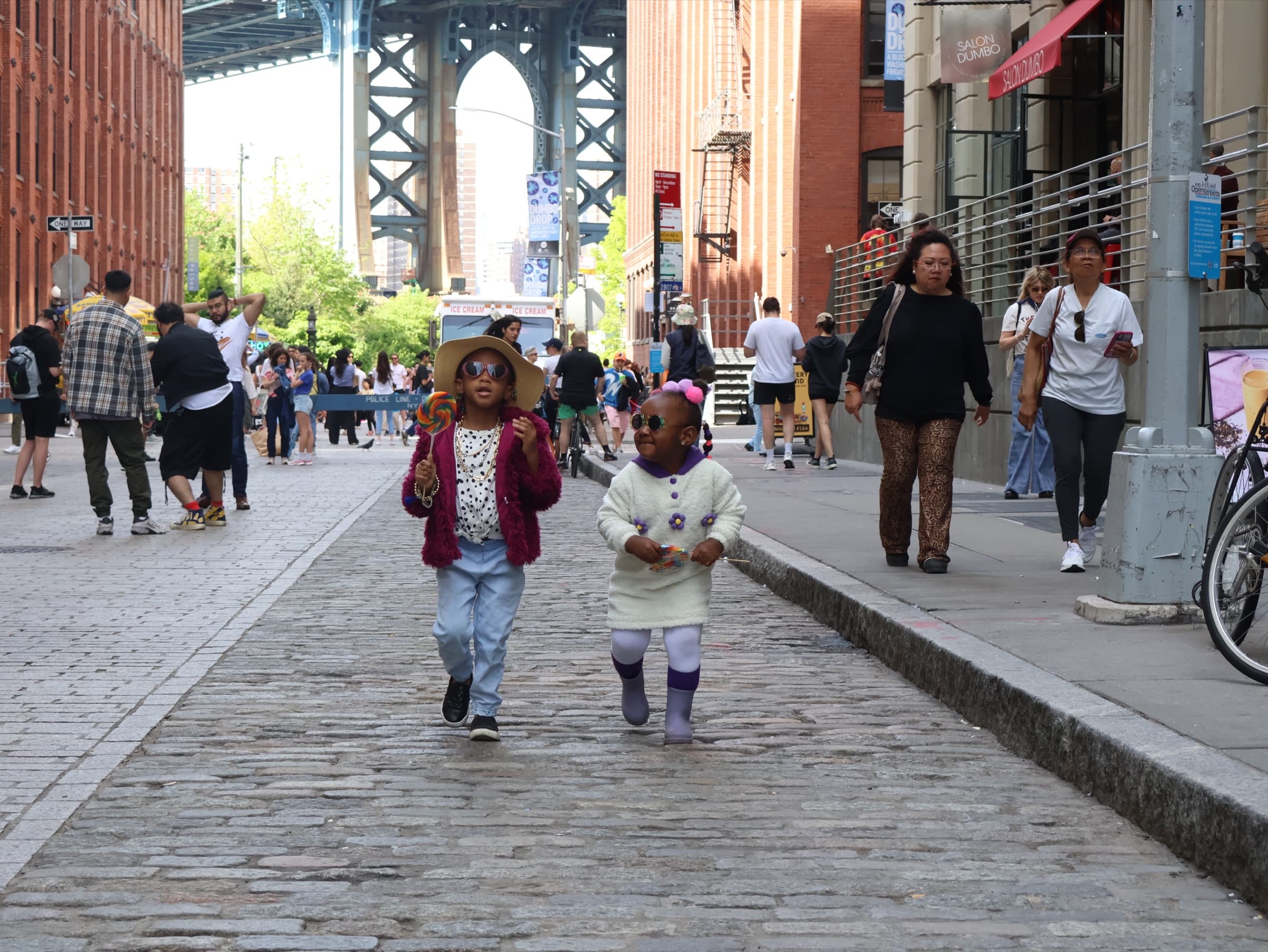 Two children in hats and sunglasses walk up a cobblestone street with the Manhattan Bridge rising behind them