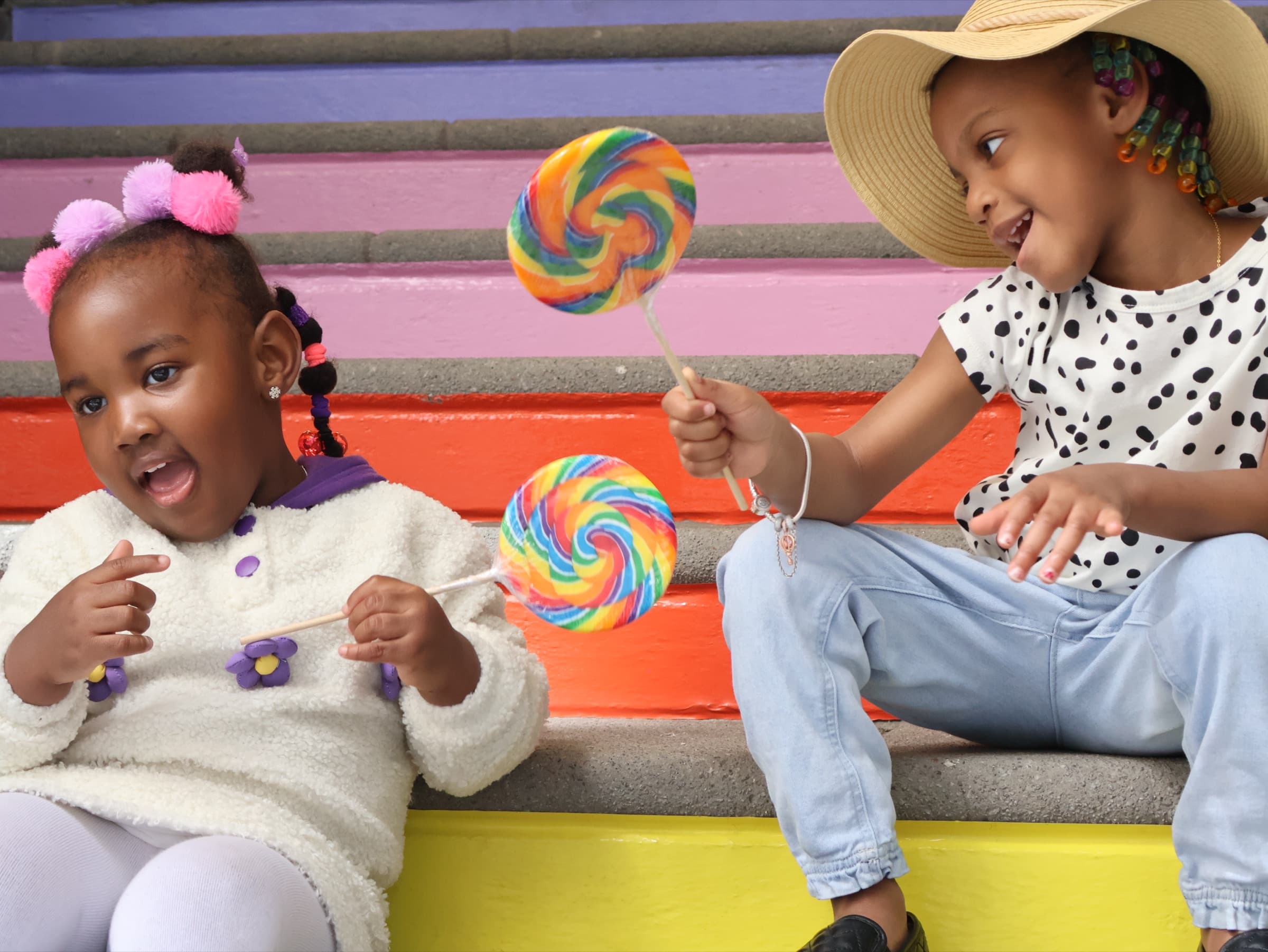 Two children sit on rainbow-painted steps holding oversized swirl lollipops, turned toward each other mid-laugh