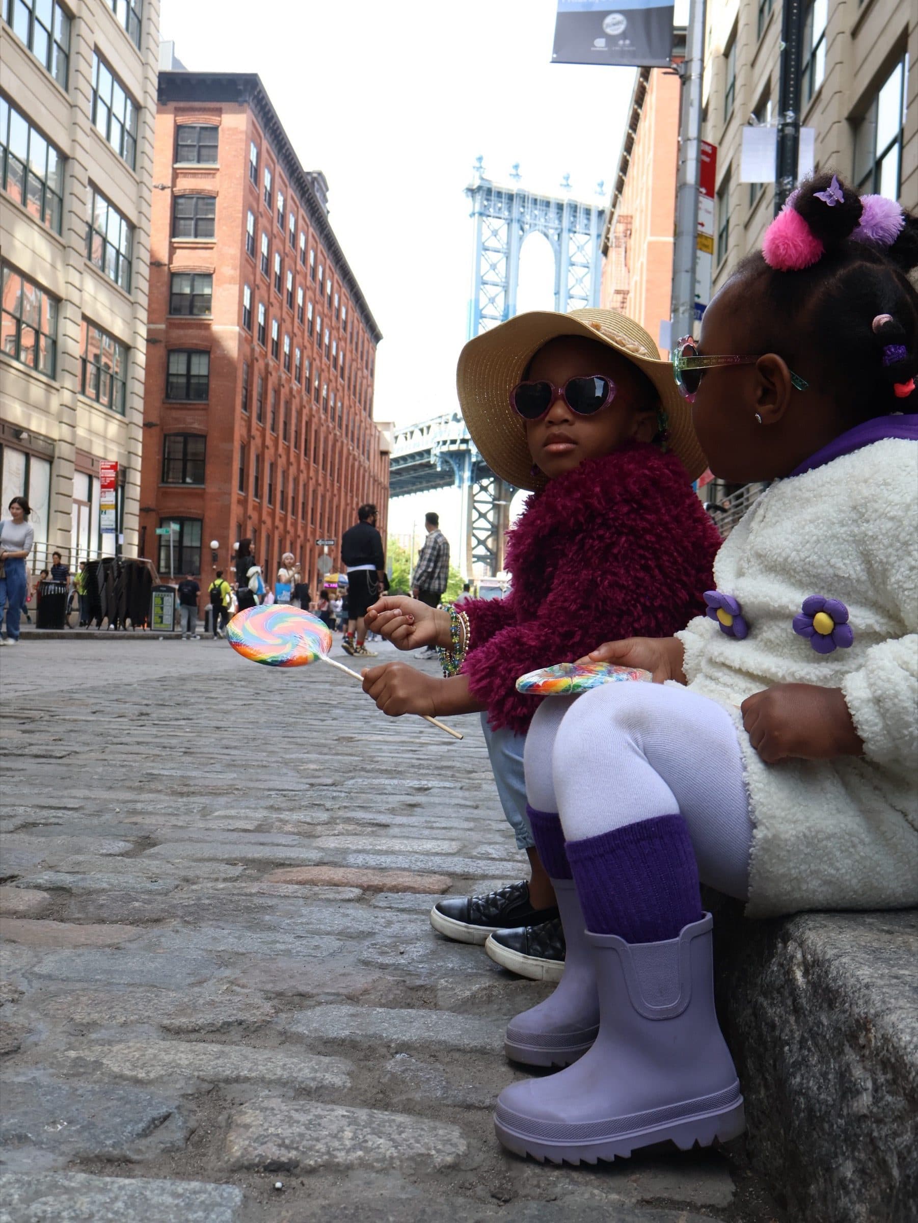 Child seated on a stone curb with a lollipop while a second child stands in the foreground, the bridge and street behind