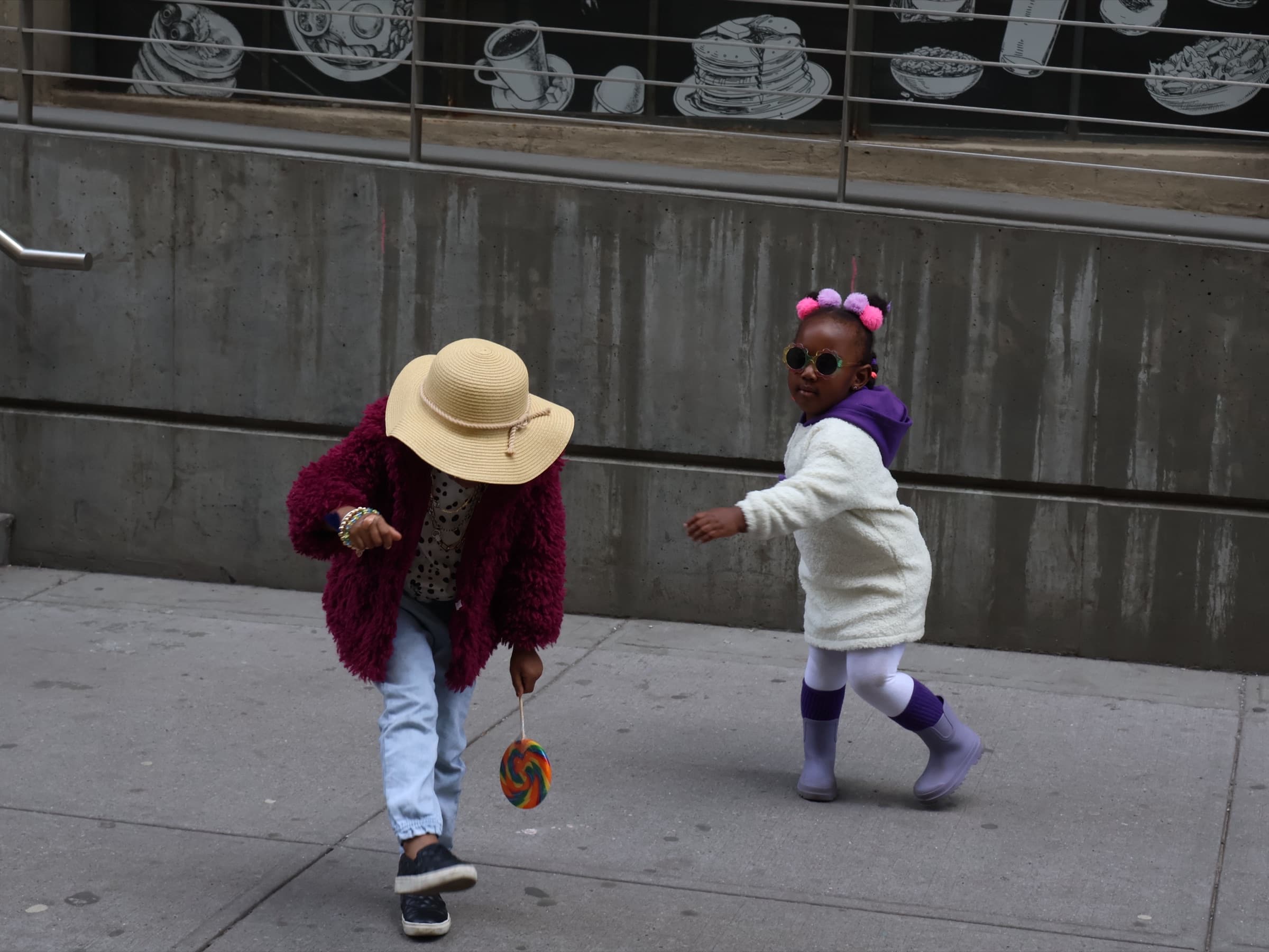 Two children dance on a city sidewalk, one bending forward under a straw sun hat while holding a lollipop