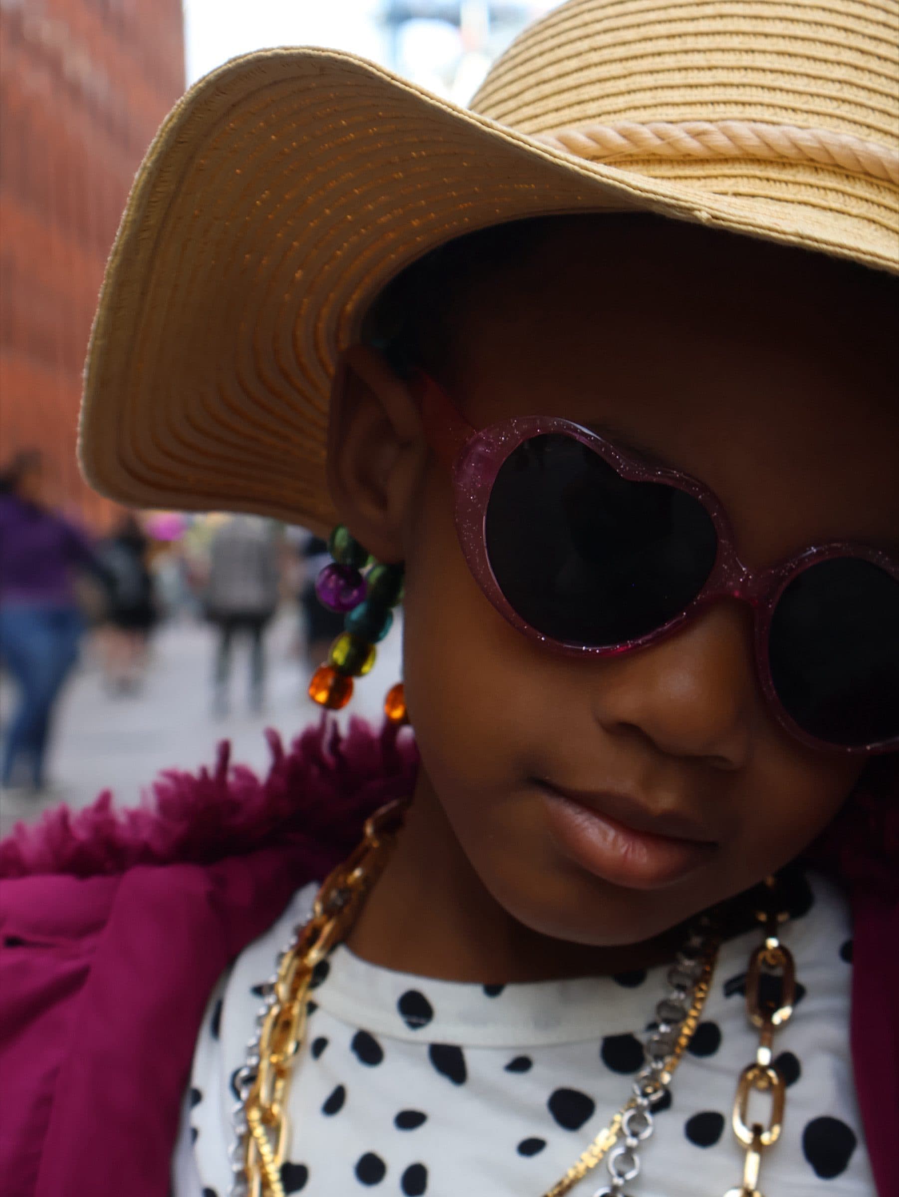 Close view of a child's face beneath a straw hat brim wearing pink heart-shaped sunglasses and beaded necklaces