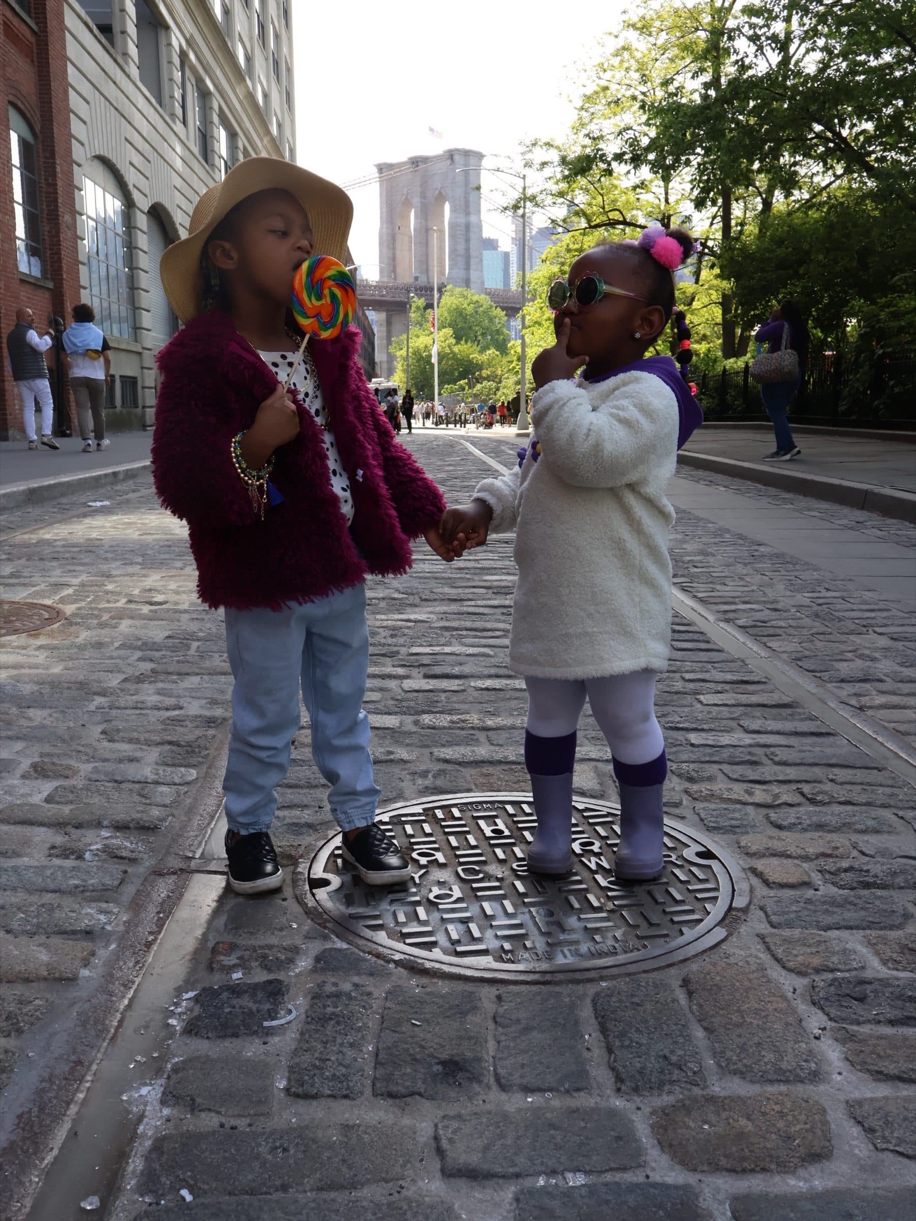 Two children stand together on an ornate manhole cover among cobblestones with city towers behind them