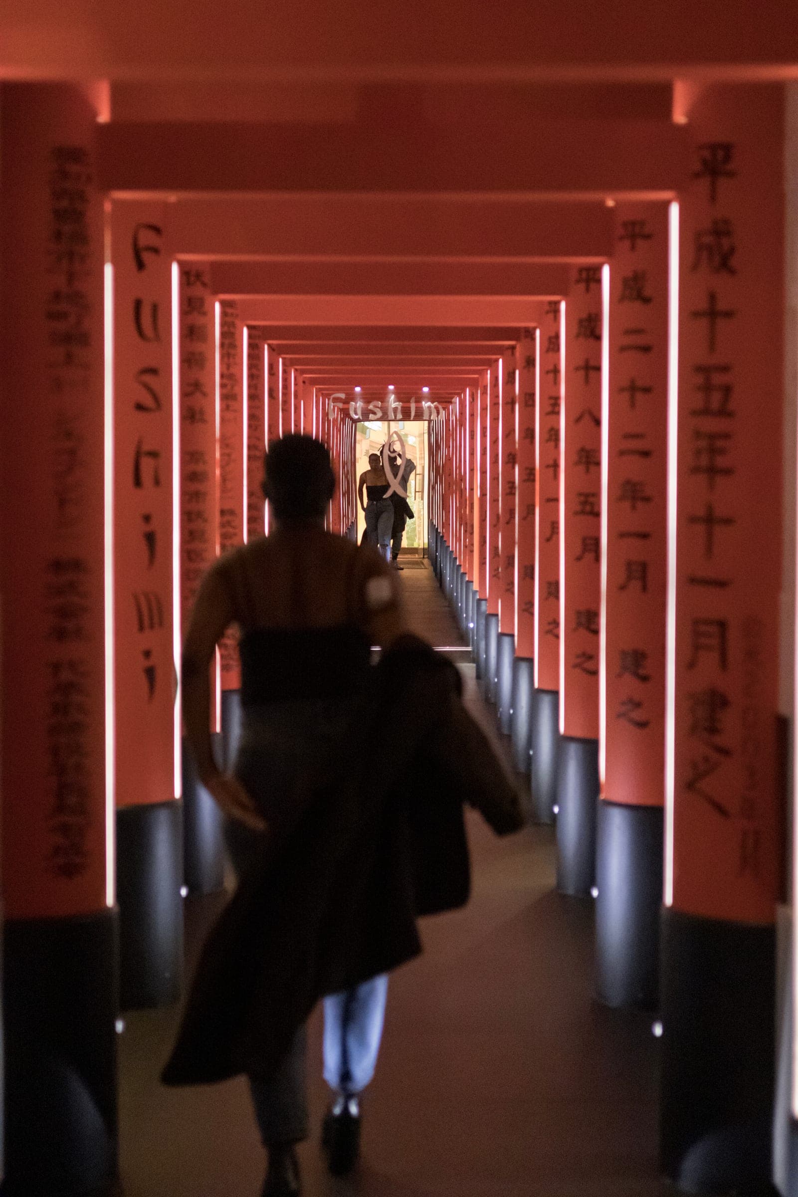 Mel walks away down a long corridor of red torii gates lit from within, a jacket over her arm