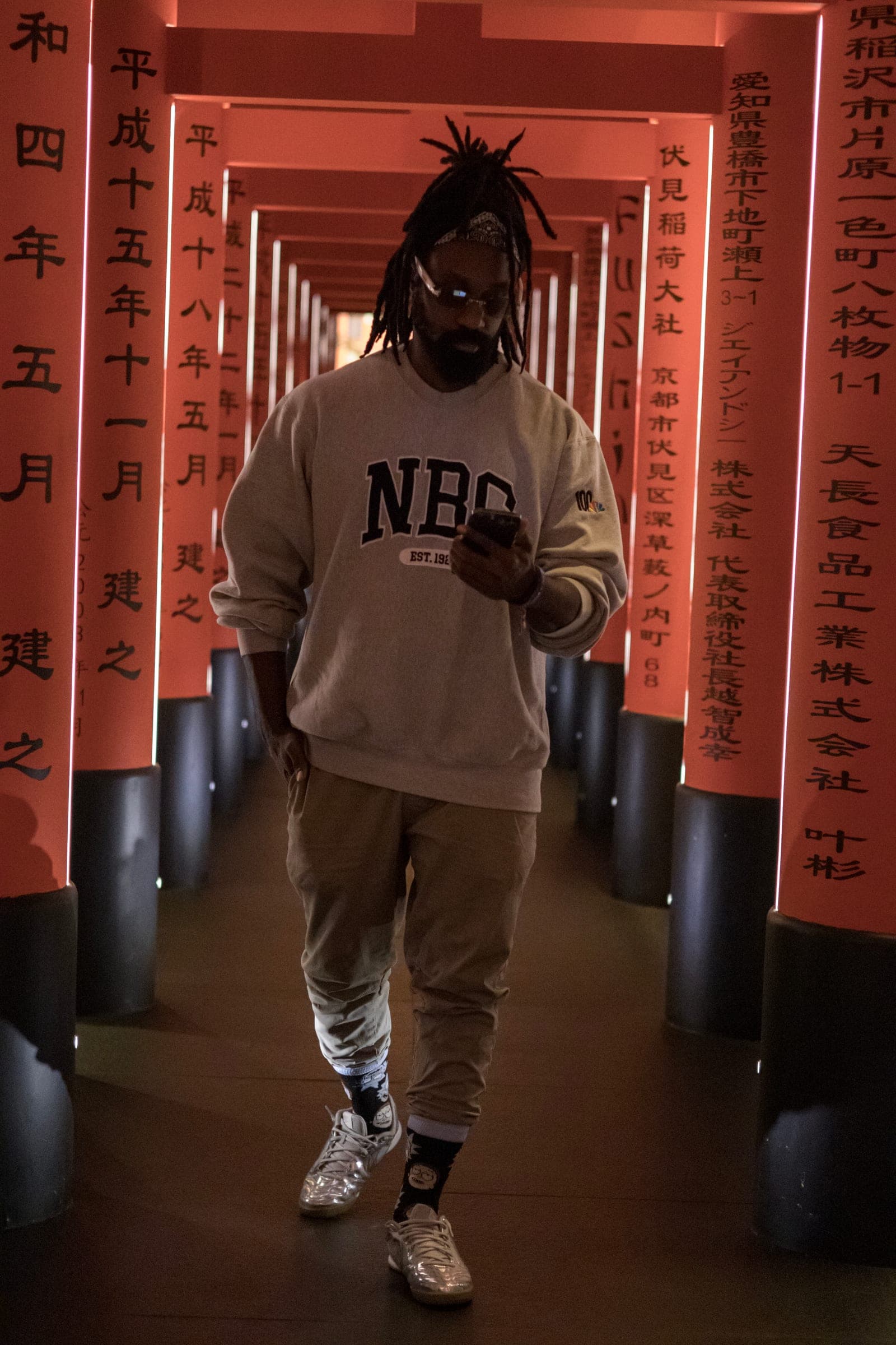 Derek walks toward the camera through the red gates, looking down at his phone