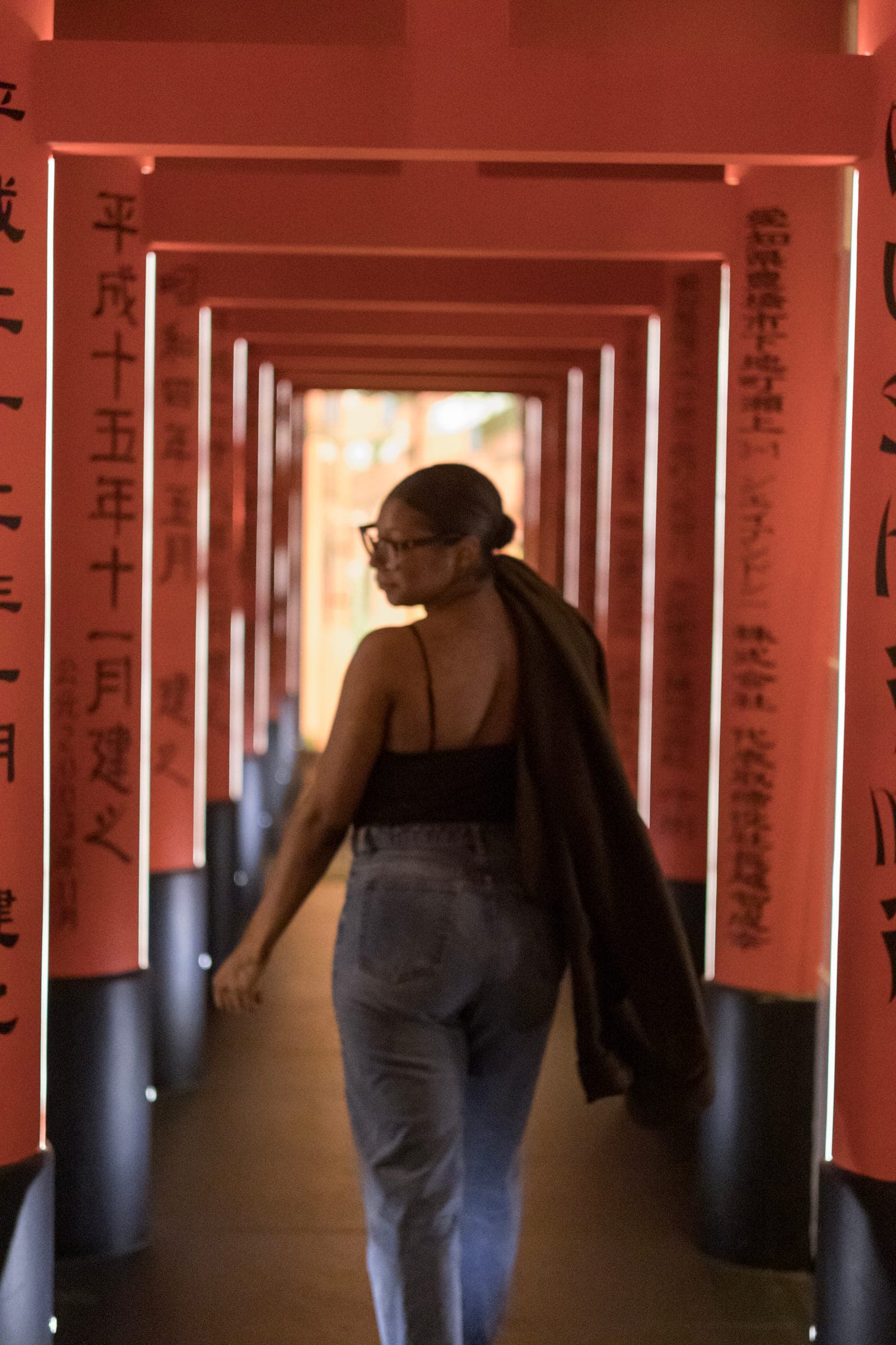 Mel walks away through the red gates, ponytail down her back, jacket slung over one shoulder