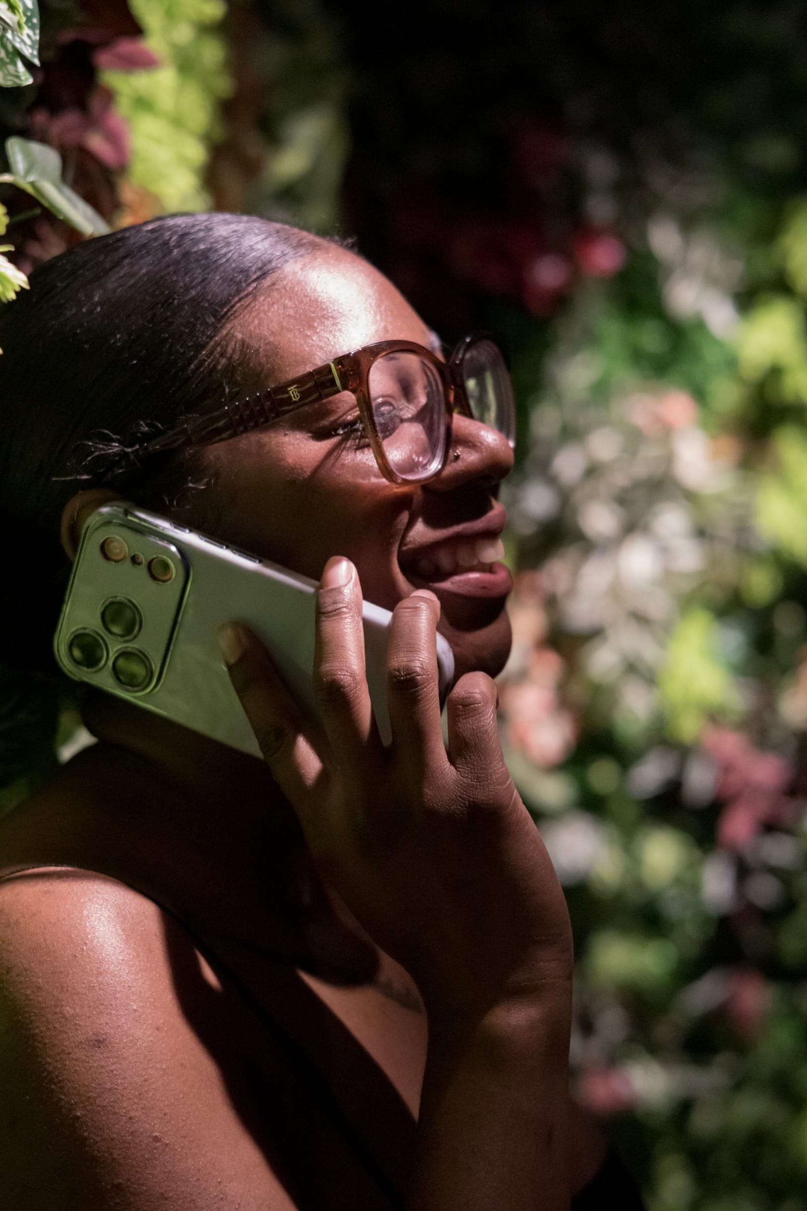 Mel laughs on a phone call, lit against a dark wall of leaves and flowers
