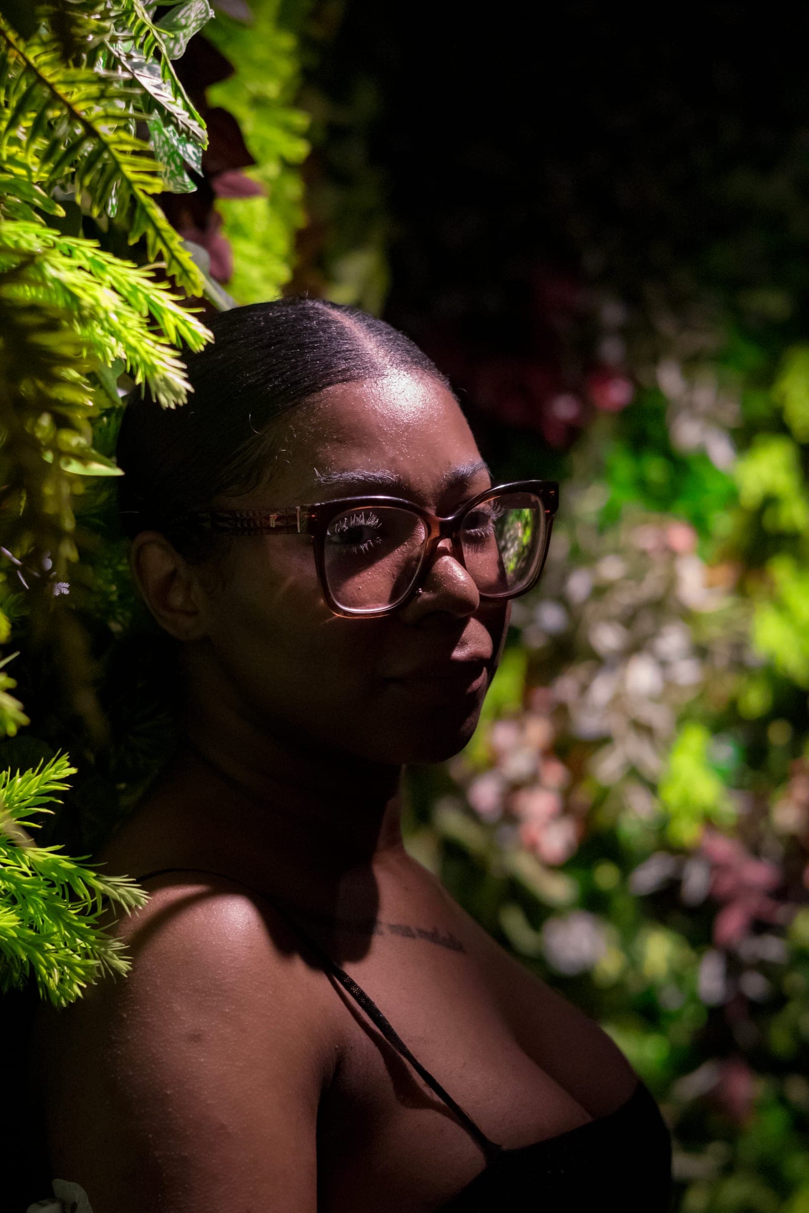 Mel glances back through glasses, framed by leaves and warm low light