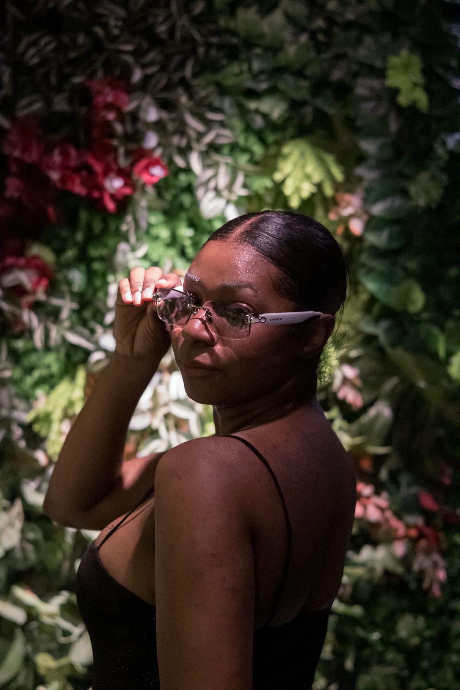 Mel adjusts her glasses and looks back over her shoulder in front of a wall of leaves and red flowers