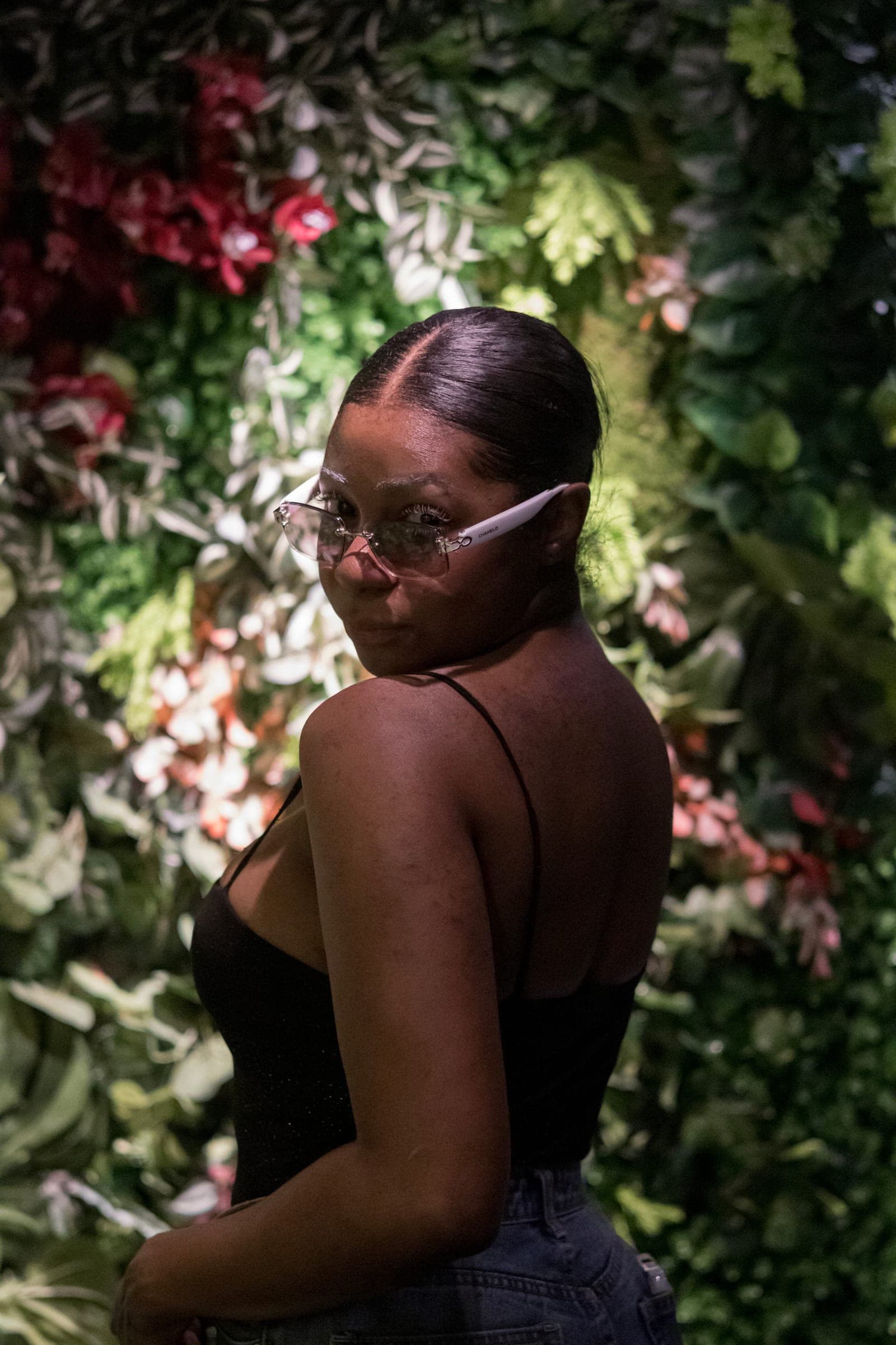 Mel turns over her shoulder toward the camera, glasses on, the flower wall bright behind her