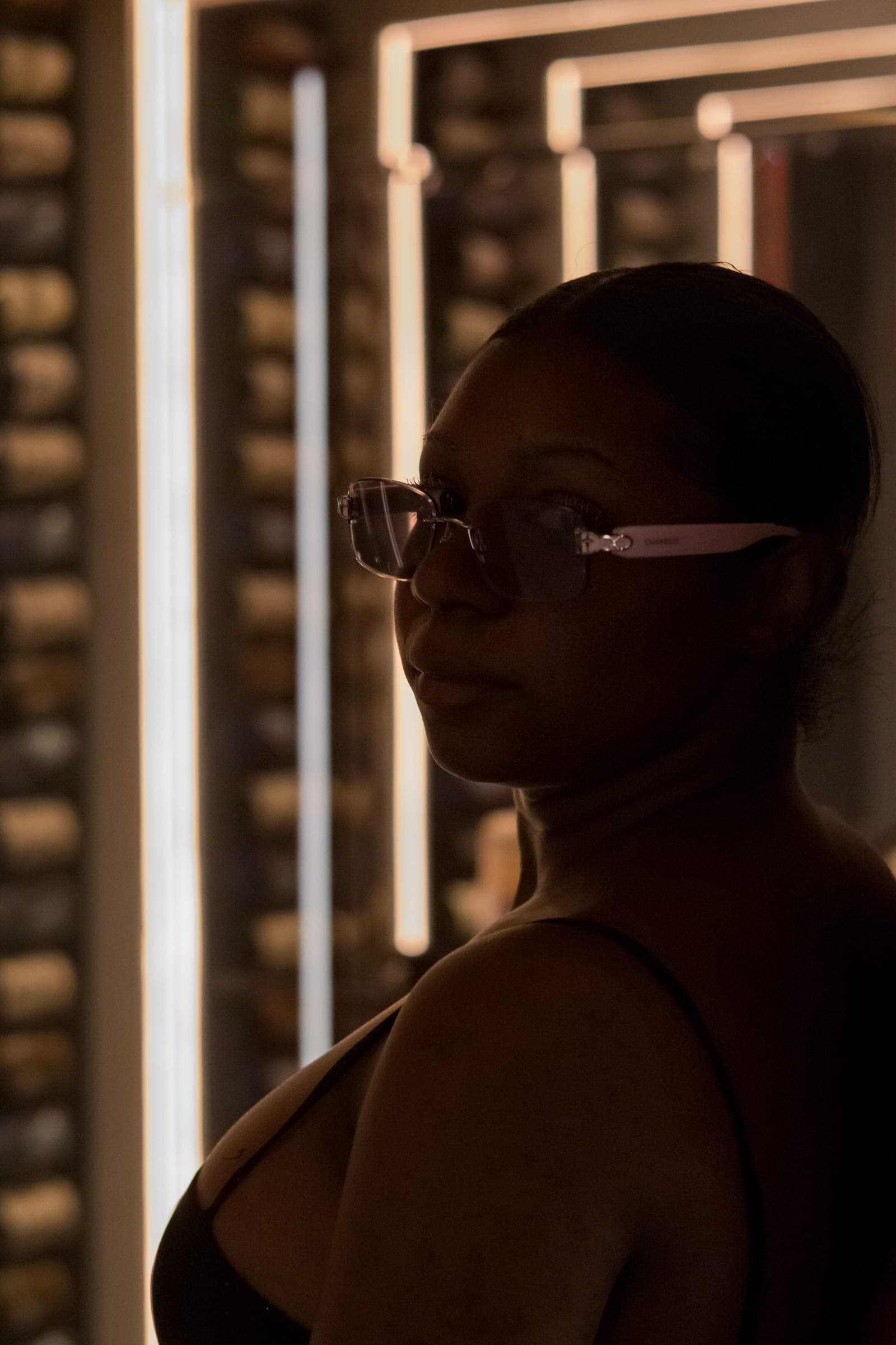 Mel stands between warm vertical light bars, half her face in shadow