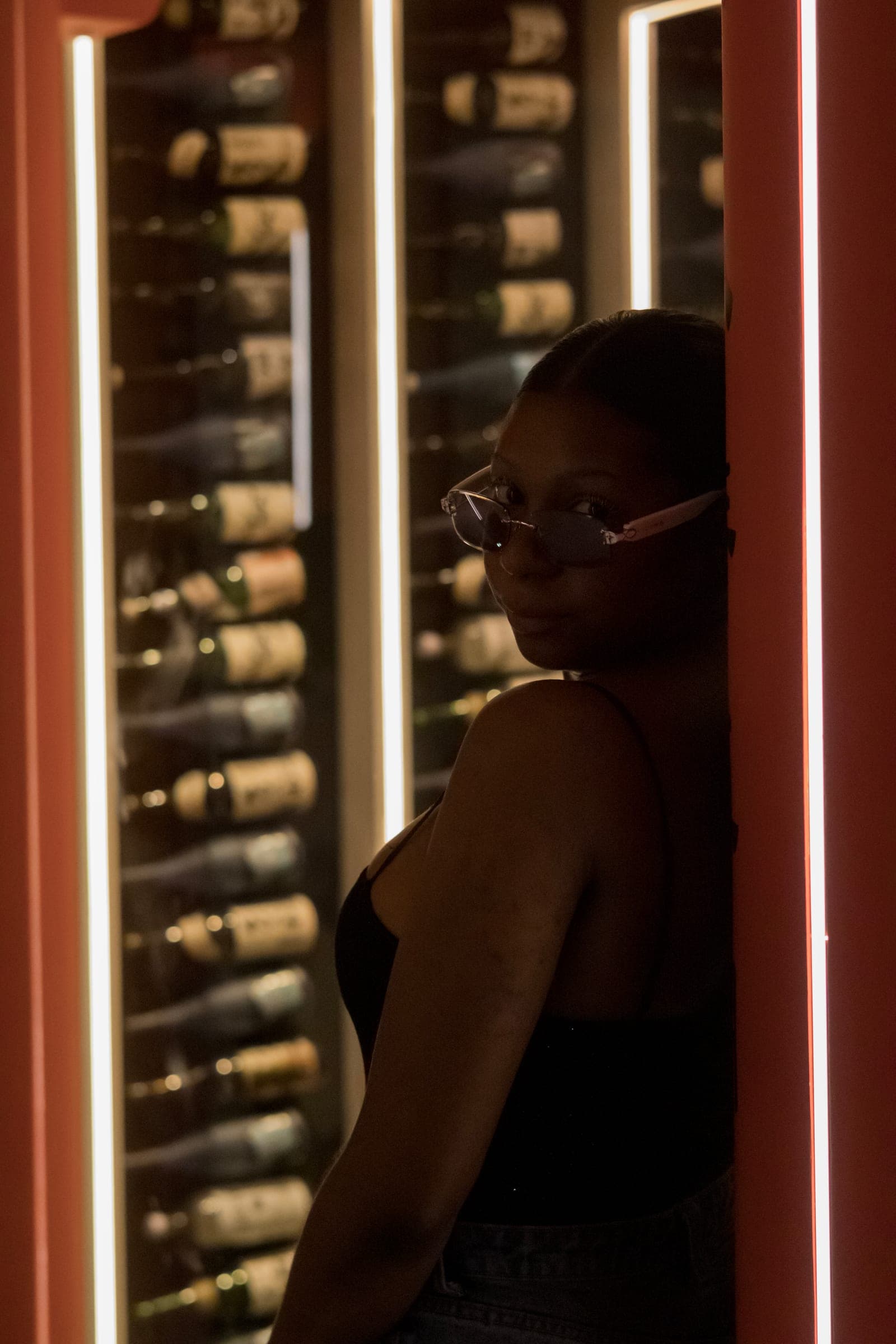Mel leans against a red pillar beside glowing light bars, looking back at the lens