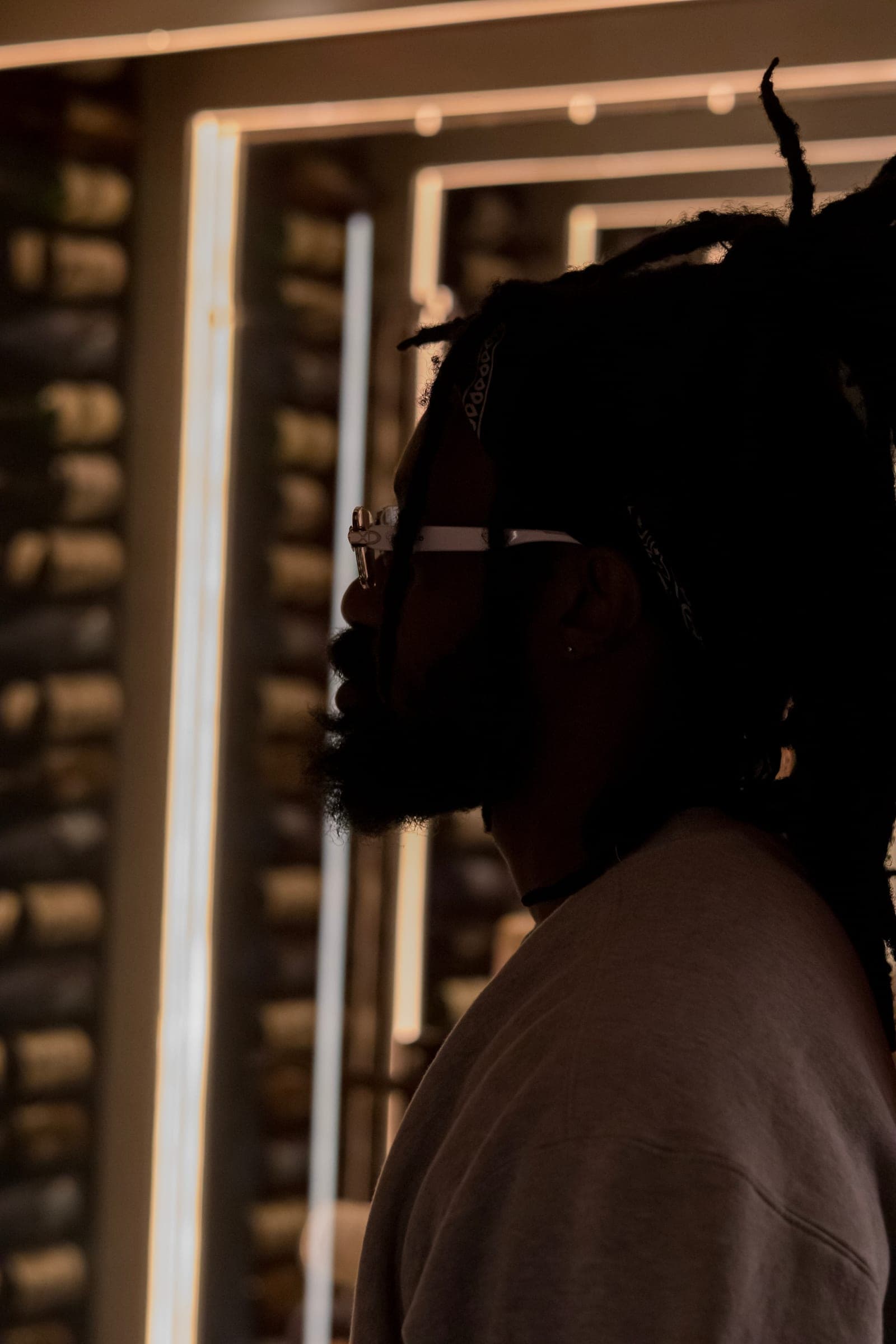 Derek in near silhouette against the light bars, locs and glasses catching the edge of the light