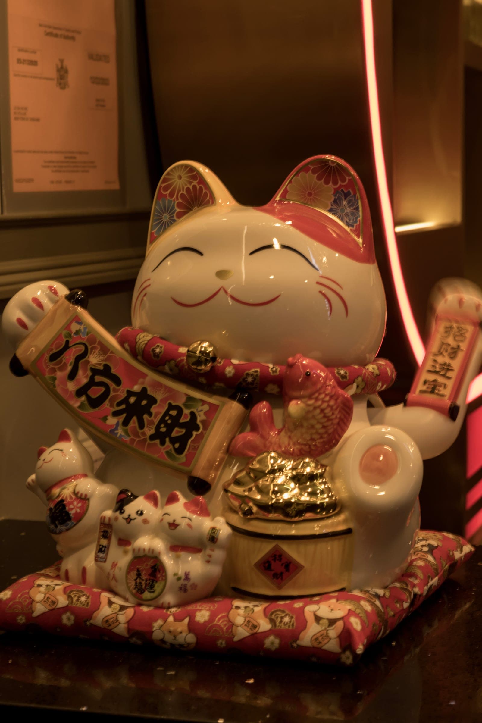 A ceramic beckoning cat with a gold coin and red cushion on a shelf, lit warm