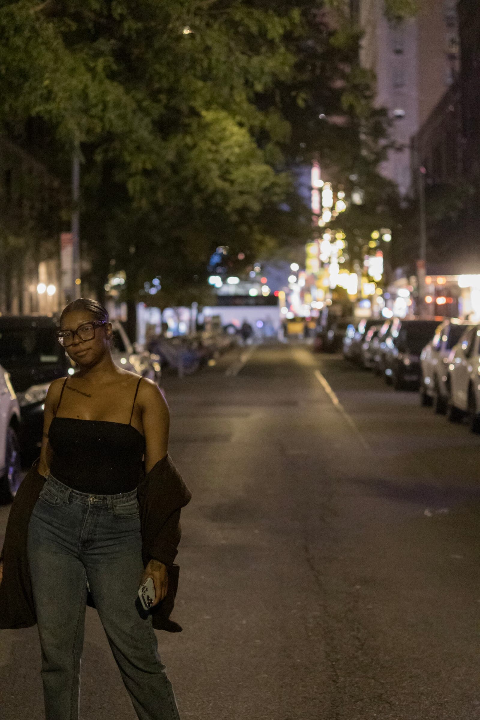 Mel walks down a Midtown street at night, jacket over her arm, parked cars and city lights behind her