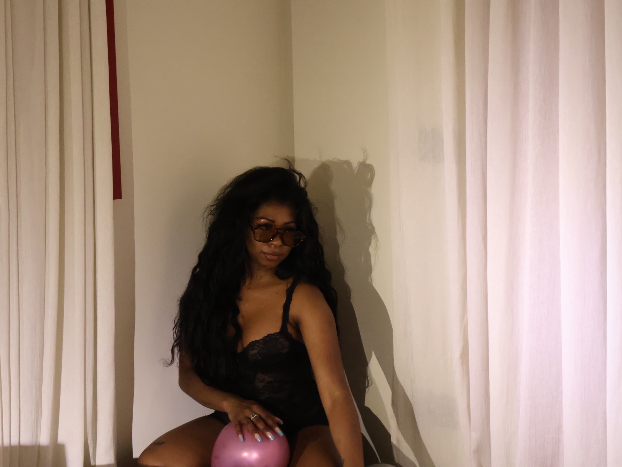 Woman in a lace bodysuit sits composed in warm lamplight, sunglasses low on her nose, one hand resting on a pink balloon