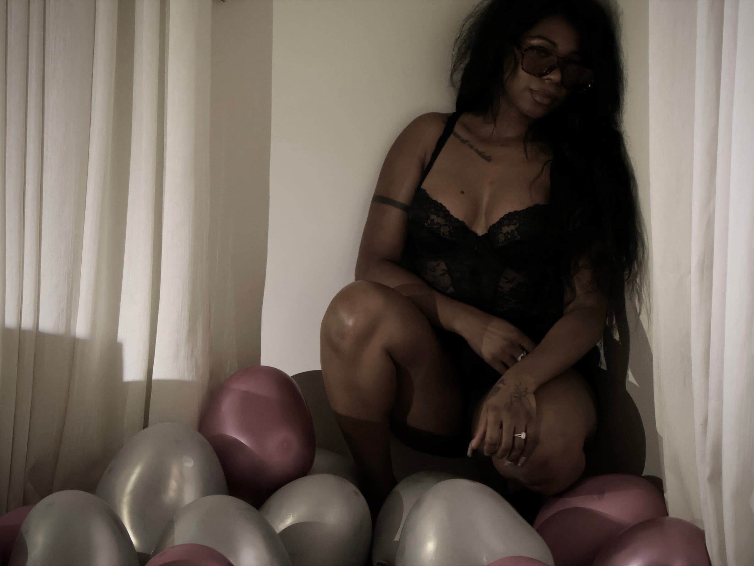 Woman sits in a curtained corner surrounded by heart-shaped balloons, arms folded over her knee, in muted warm light