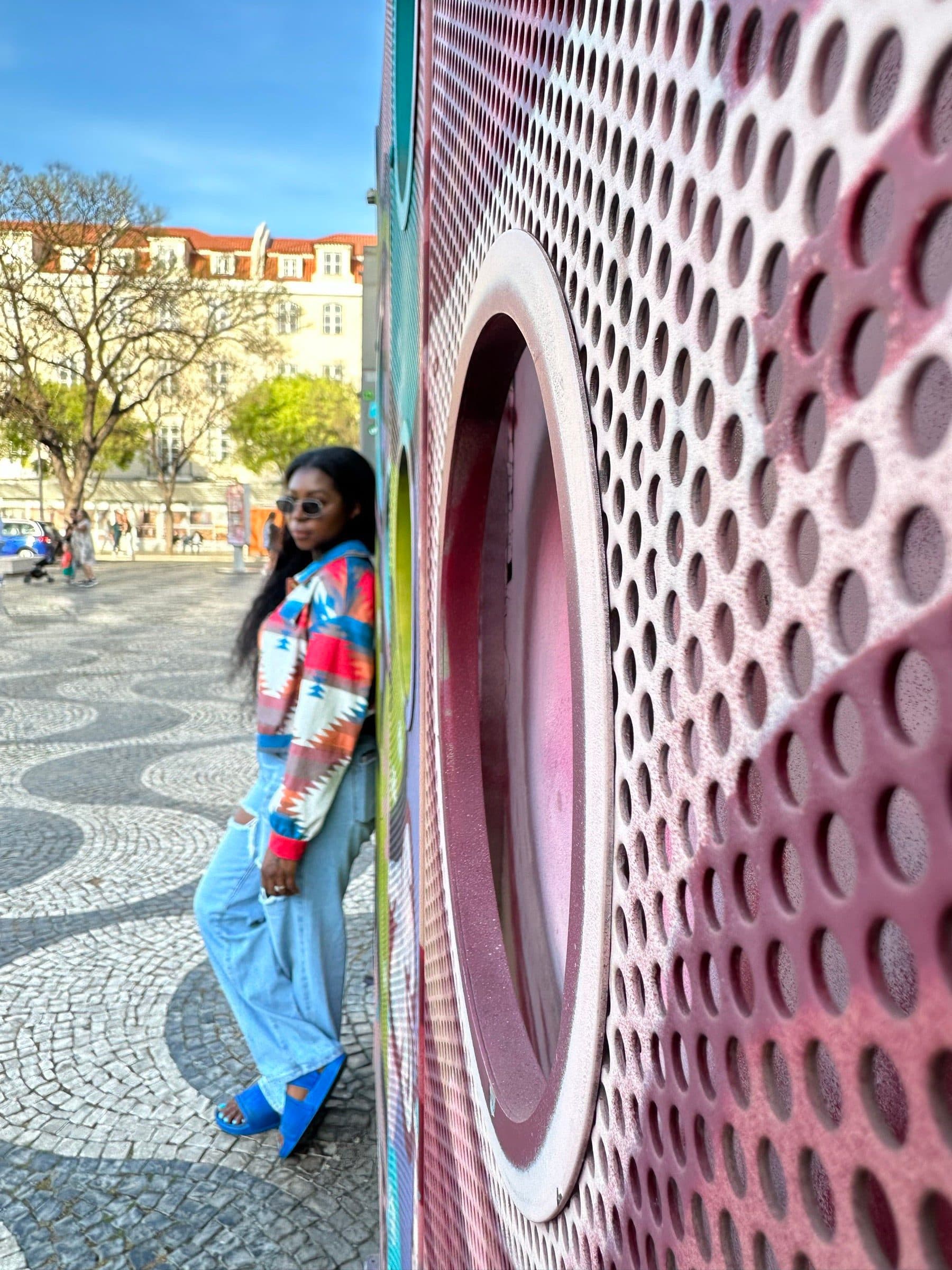 Woman leans against a large perforated pink sculpture on wavy black-and-white mosaic pavement in a European square
