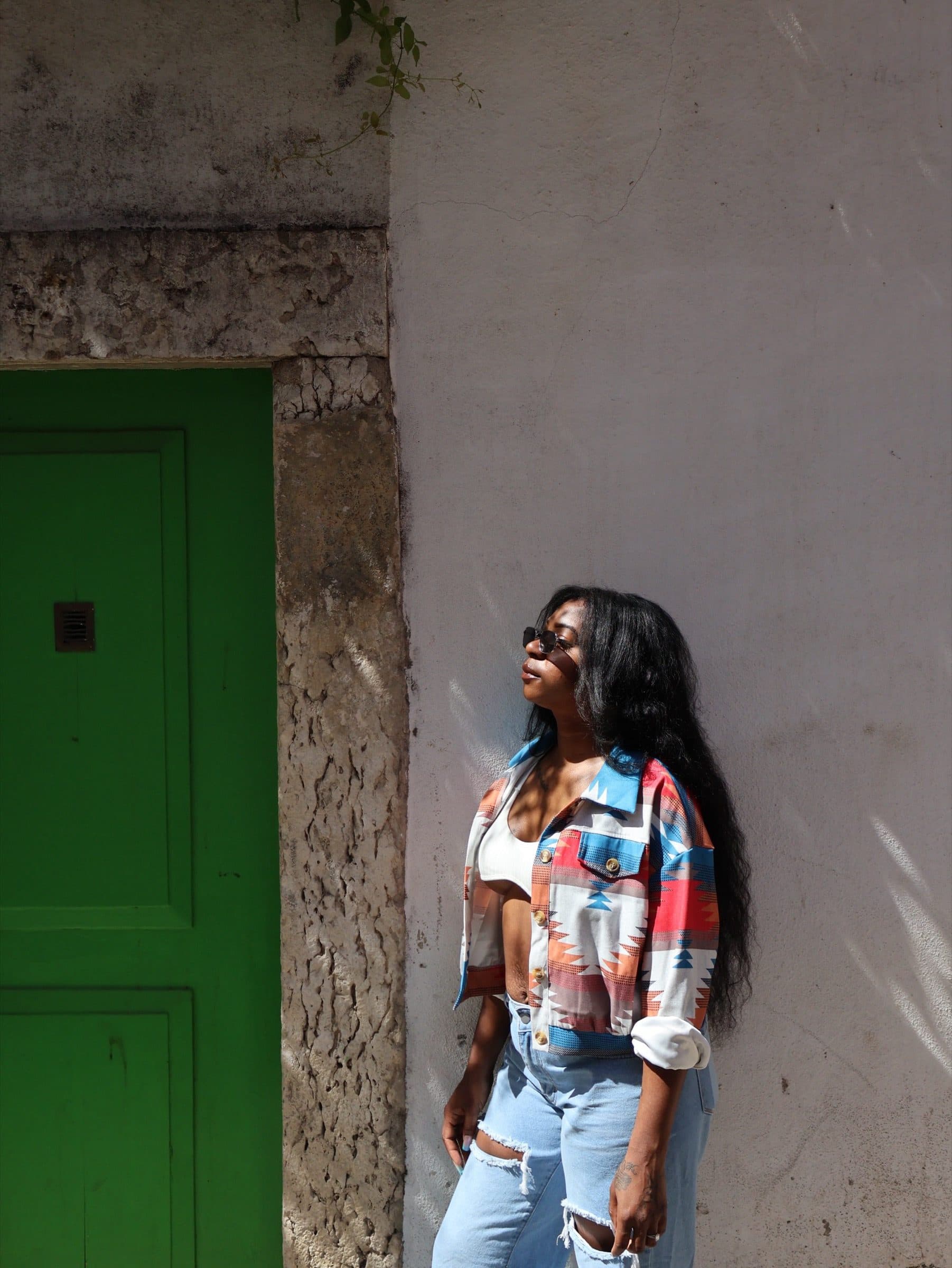 Woman in an Aztec-print jacket and jeans leans against a weathered plaster wall beside a green wooden door in hard sunlight