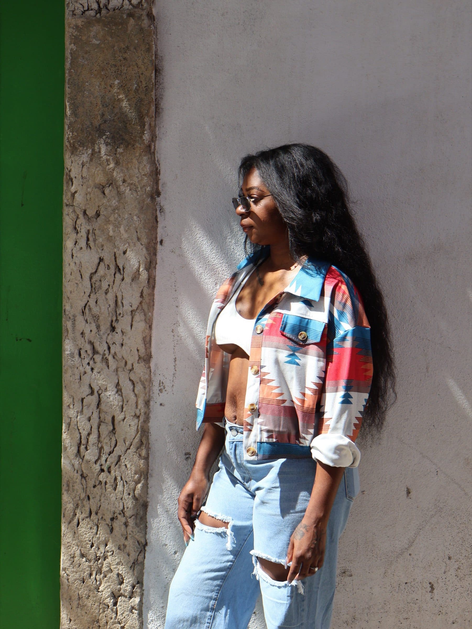 Woman leans back on an aged stone-and-plaster wall in bright sun, dark hair falling over an Aztec-print jacket