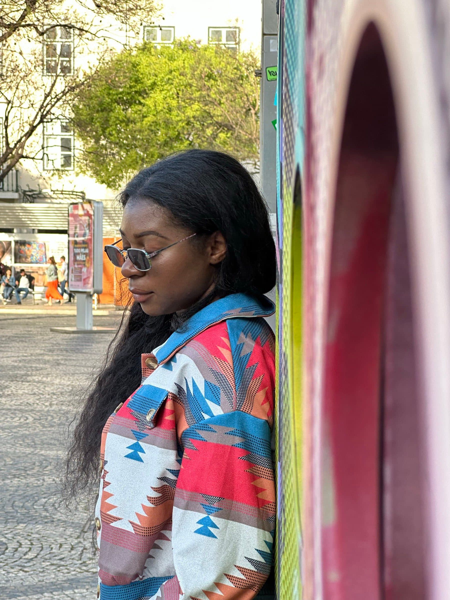 Woman in an Aztec-print jacket glances down while walking beneath a colorful canopy on a European shopping street