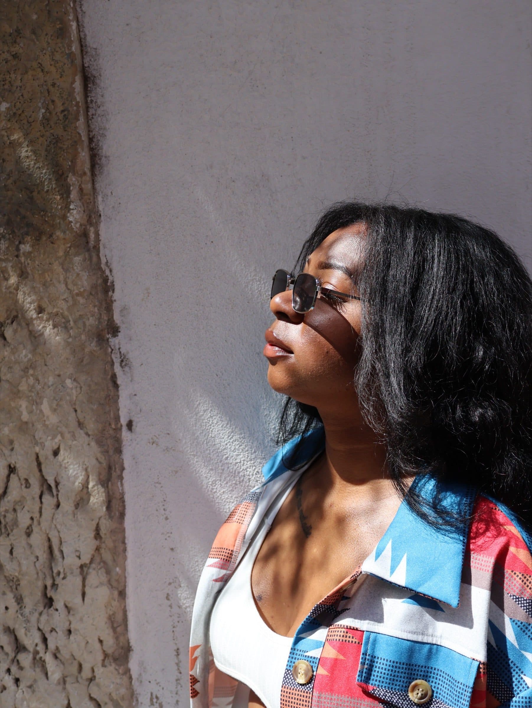 Close portrait of a woman with head tilted back against a sunlit plaster wall, holding sunglasses near her chin