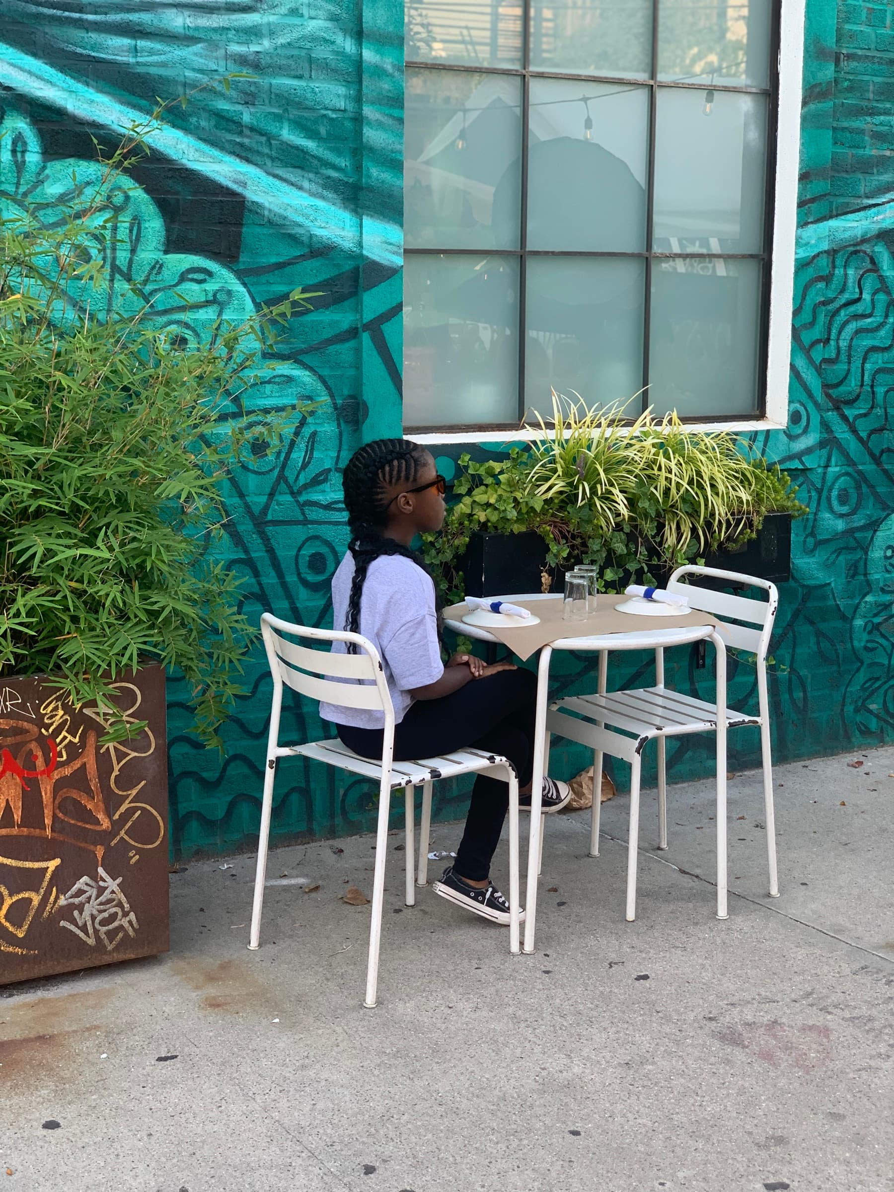 Child sits alone at a small white café table on a sidewalk beside a teal patterned mural and green planters
