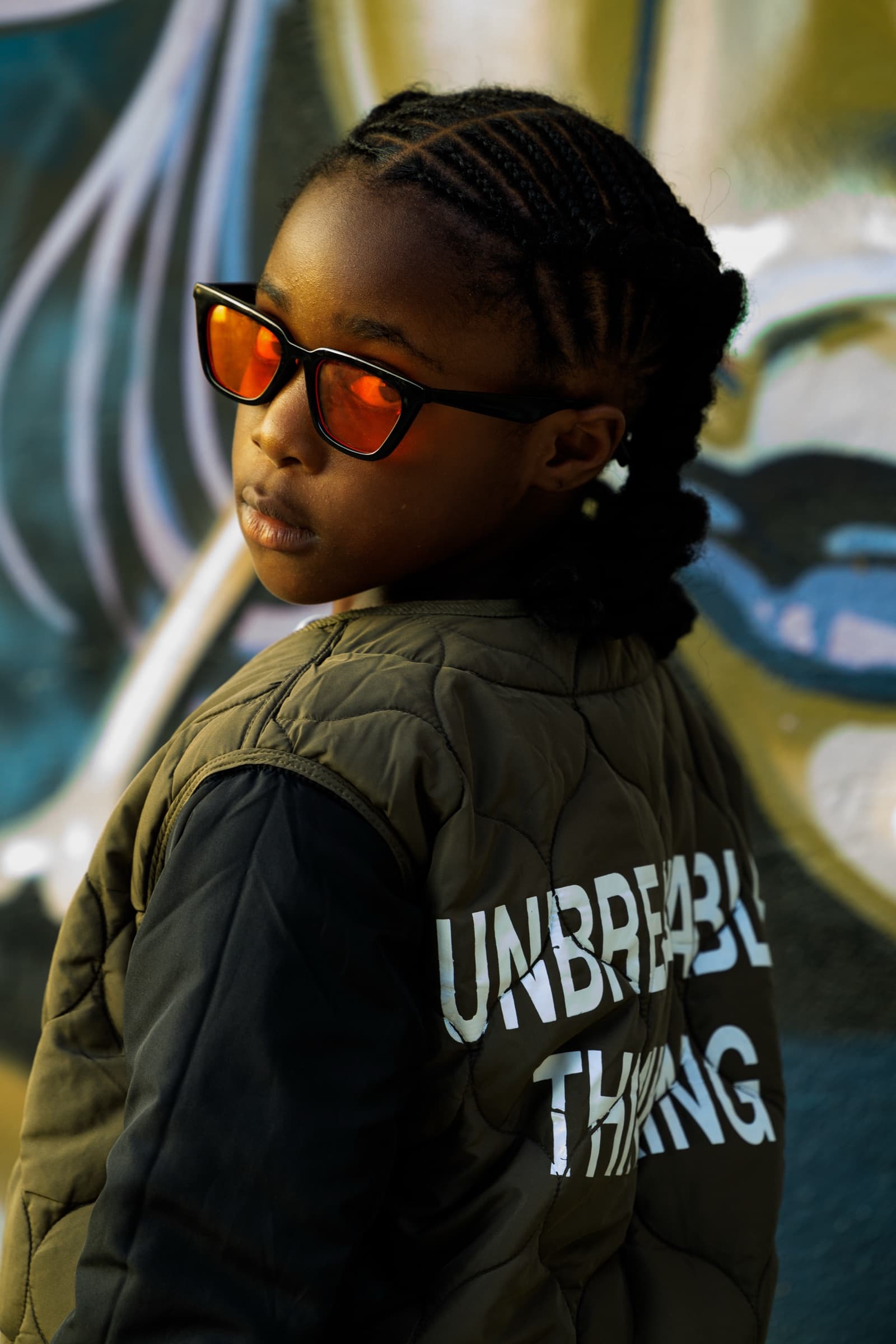 Child in an olive quilted jacket printed 'Unbreakable Thinking' looks over her shoulder wearing orange-tinted sunglasses in front of a painted mural