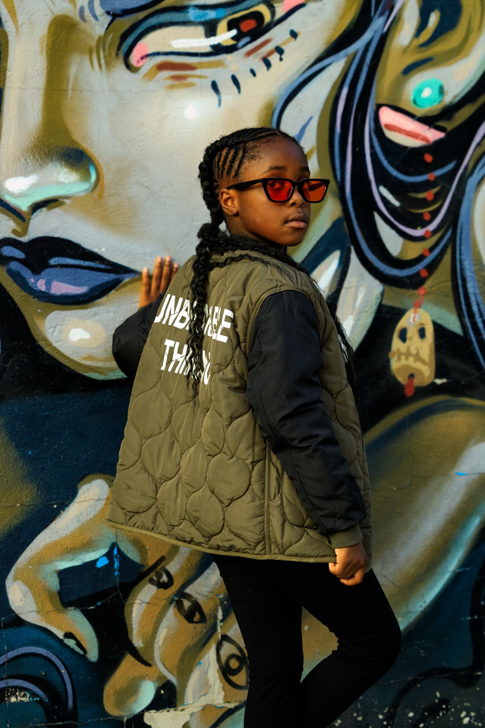 Child in red sunglasses and an olive quilted jacket stands with hand on hip before a large mural of a painted face