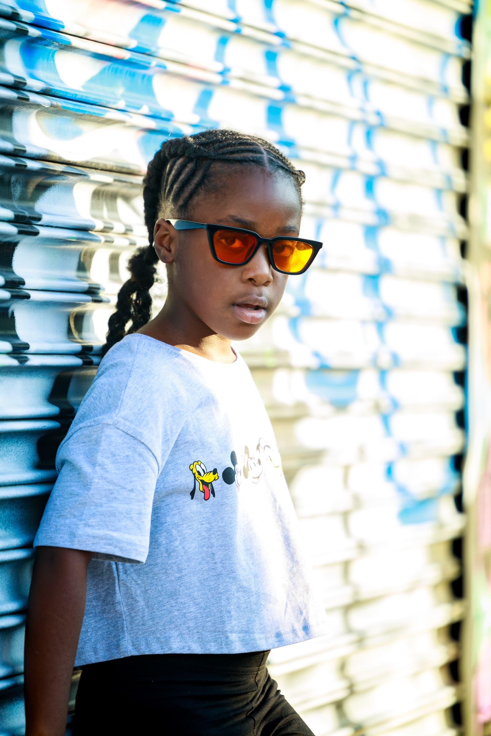 Child in orange-tinted sunglasses glances at the camera in front of a sunlit blue-and-white metal roll gate