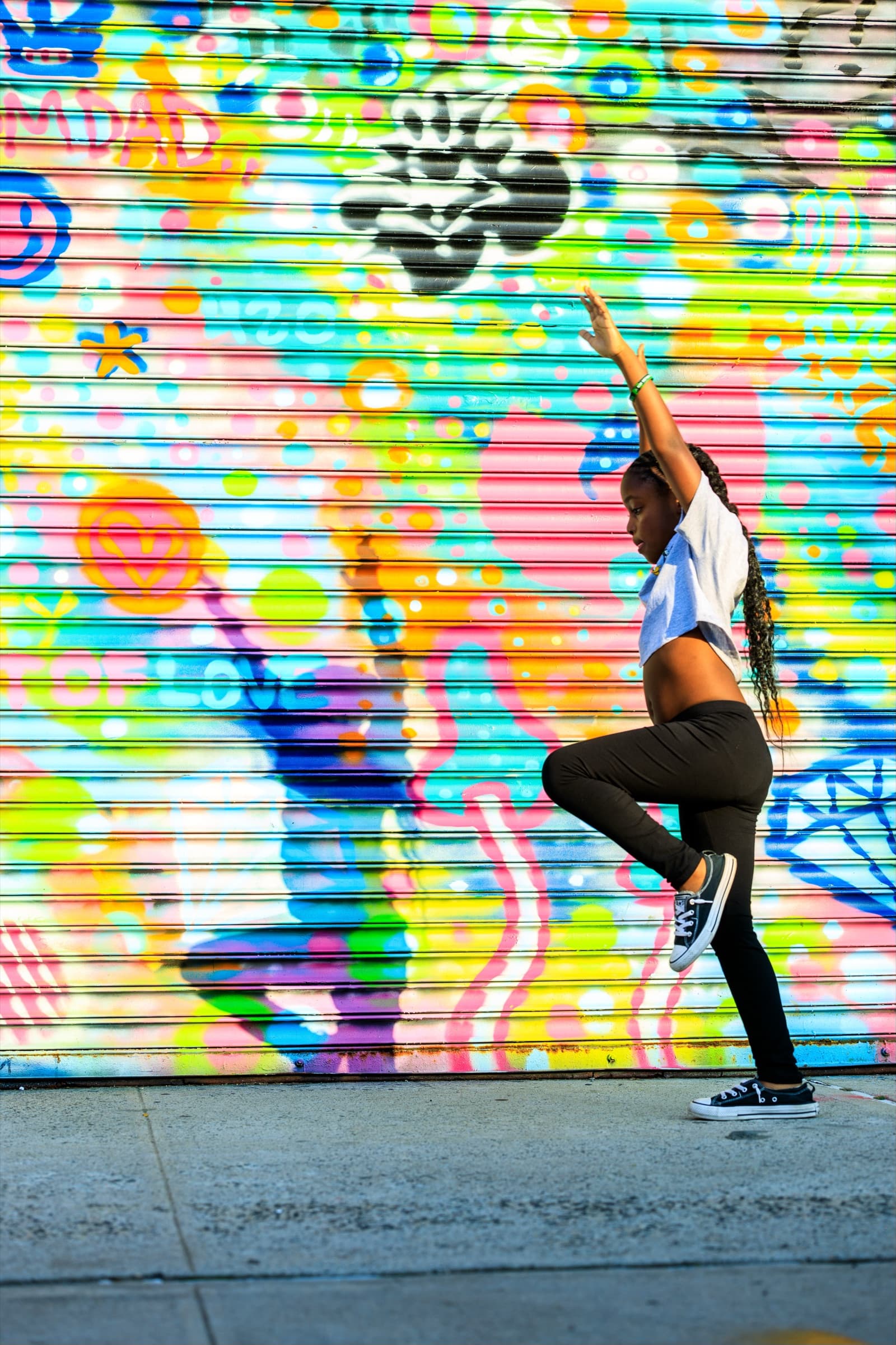 Child poses mid-movement with one arm raised in front of a brightly painted roller shutter on a city sidewalk