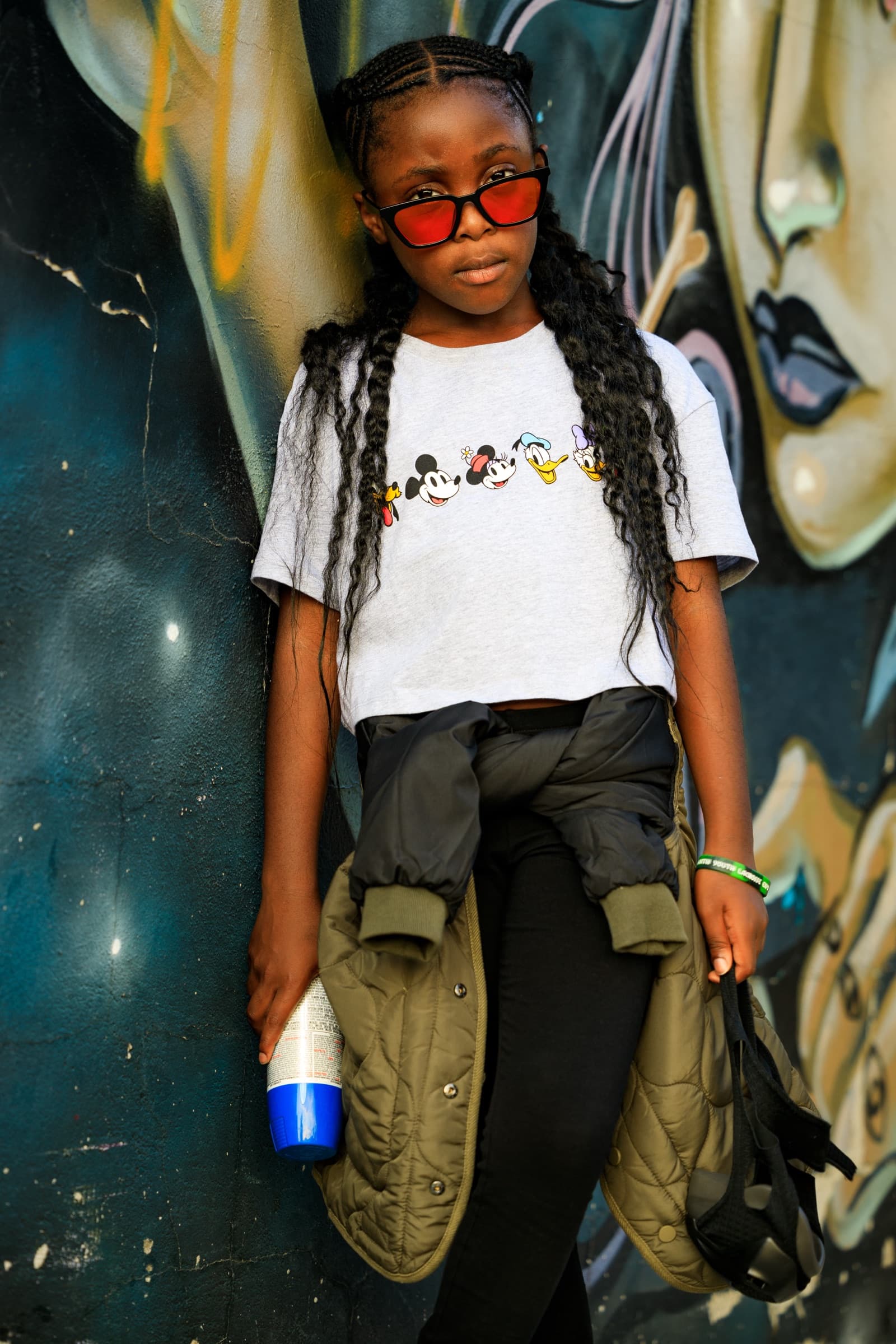 Child with long braids and red sunglasses stands facing the camera in a grey cartoon t-shirt, jacket tied at her waist, against a mural wall