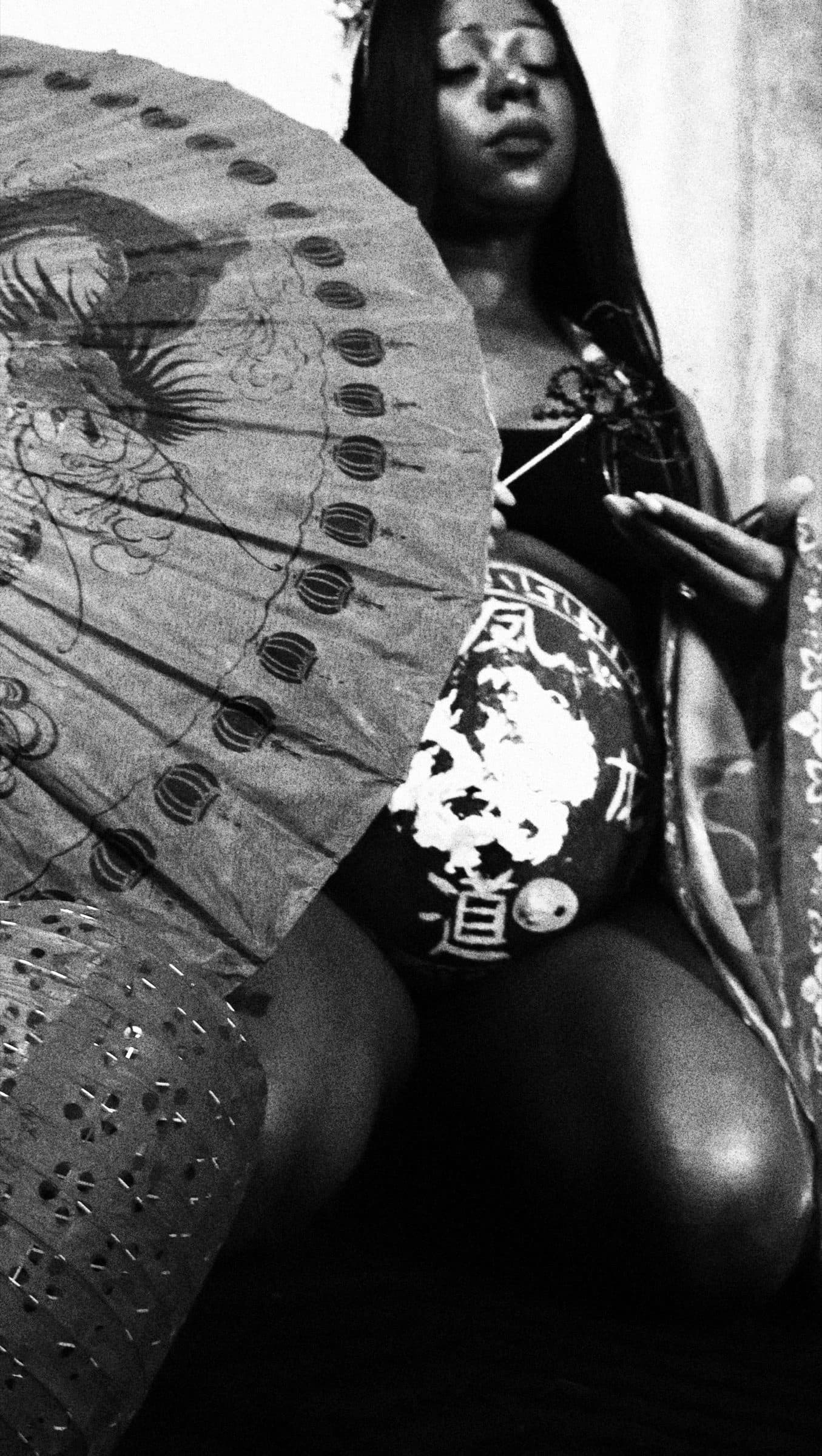 Black-and-white image of a reclining pregnant woman holding a large paper fan decorated with a dragon over her painted belly