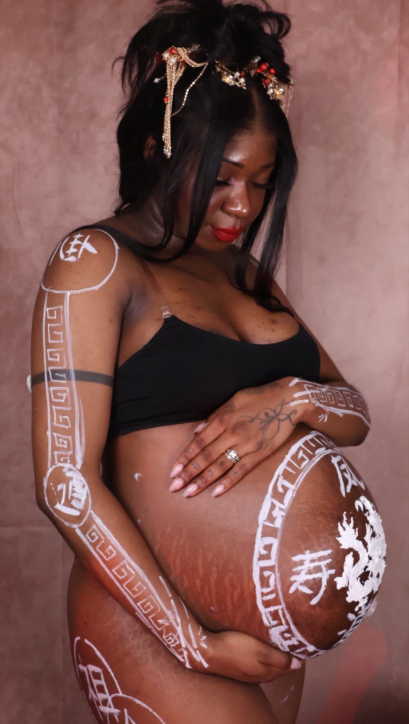 Pregnant woman reclines with white painted characters and patterned borders across her belly and arm, wearing a gold and red floral headpiece