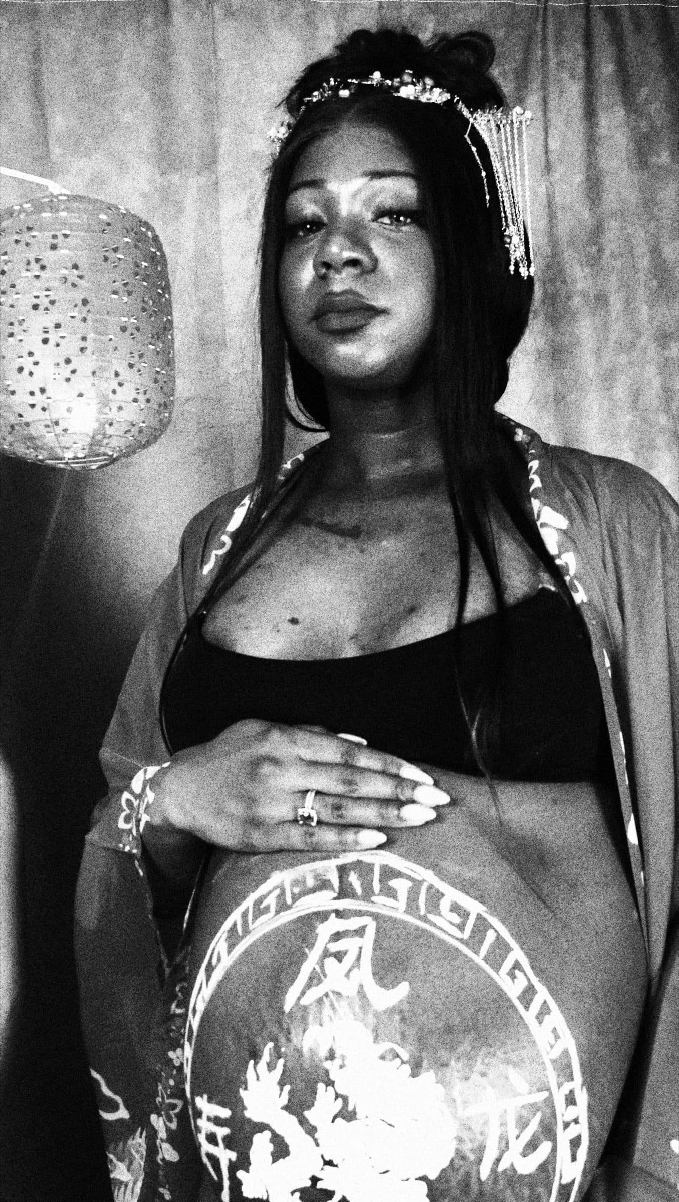 Black-and-white portrait of a pregnant woman looking at the camera, hand resting on her painted belly beneath a paper lantern