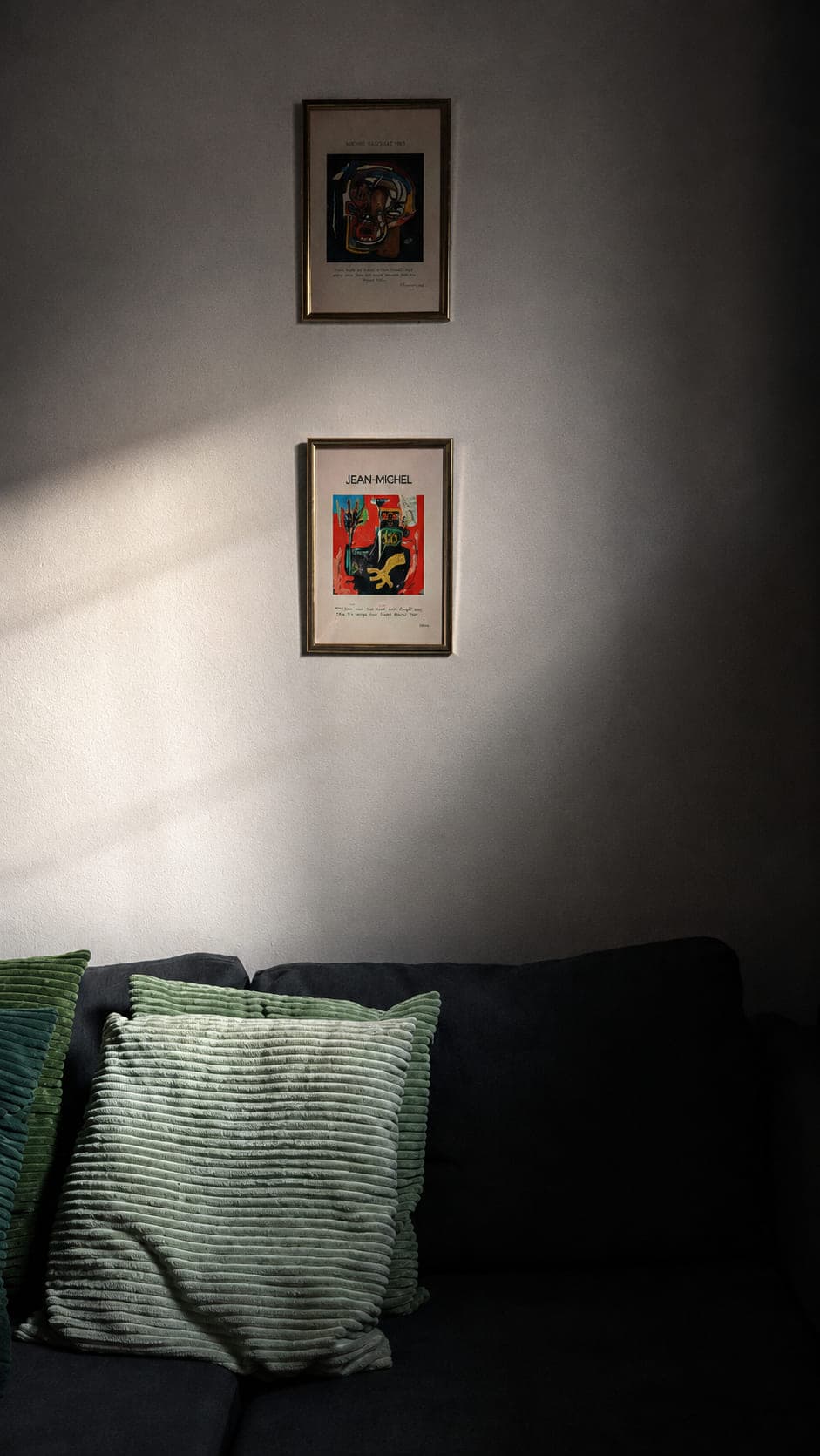Two framed prints on a wall crossed by a hard shaft of daylight, green pillows falling into deep shadow