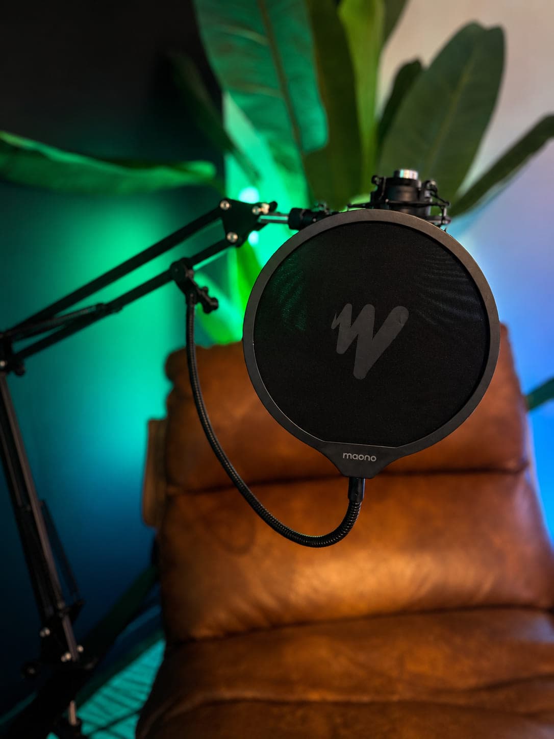 Close view of a pop filter and microphone on a boom arm above a leather chair, green and blue light behind