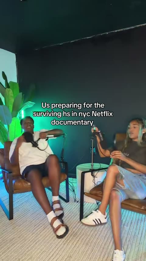 Two creators in the studio's leather chairs, microphones on boom arms, performing a comedy bit to camera under green and blue light