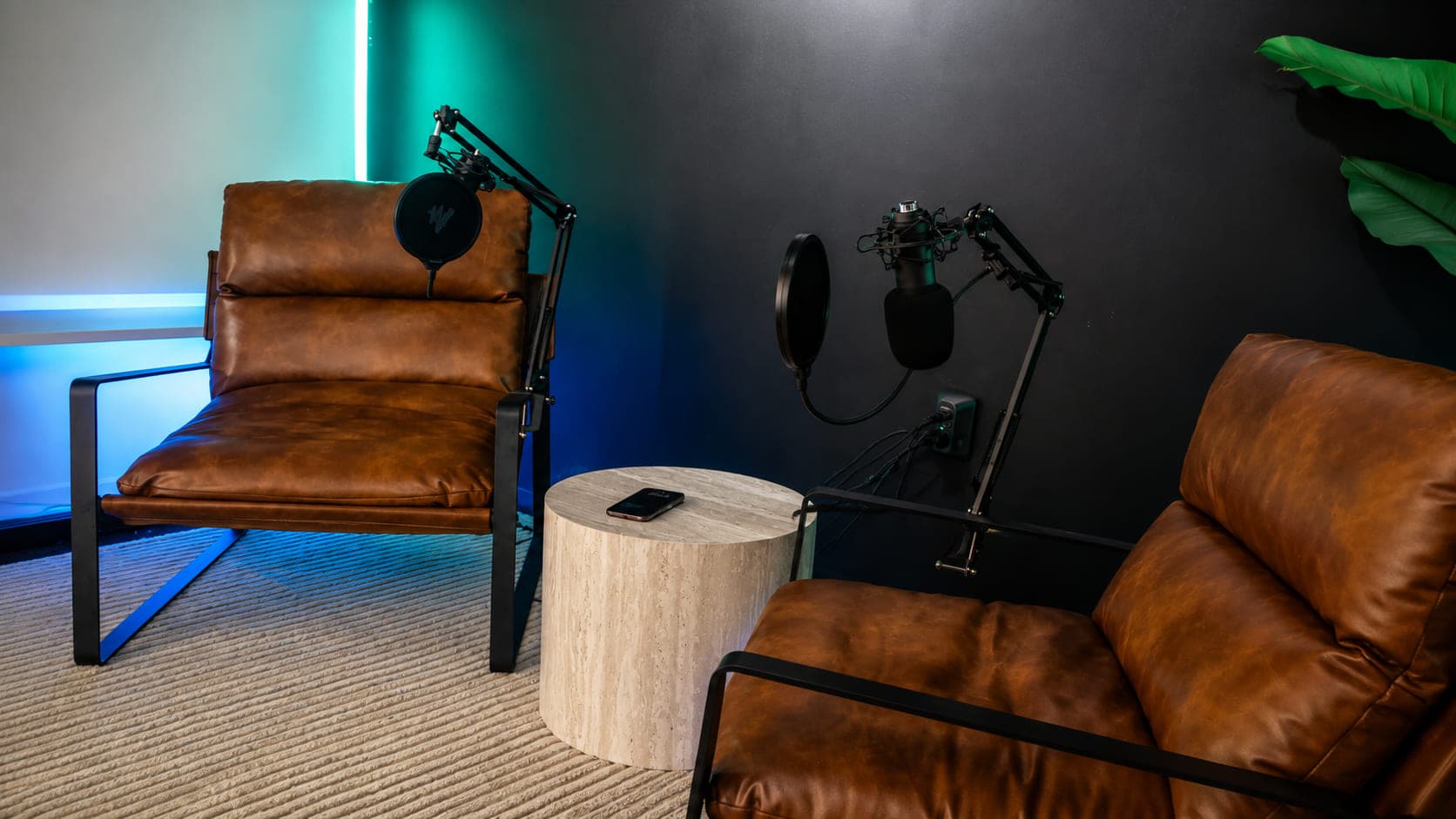 Two leather chairs facing each other with a microphone on a boom arm at each, a stone drum table between them under teal light