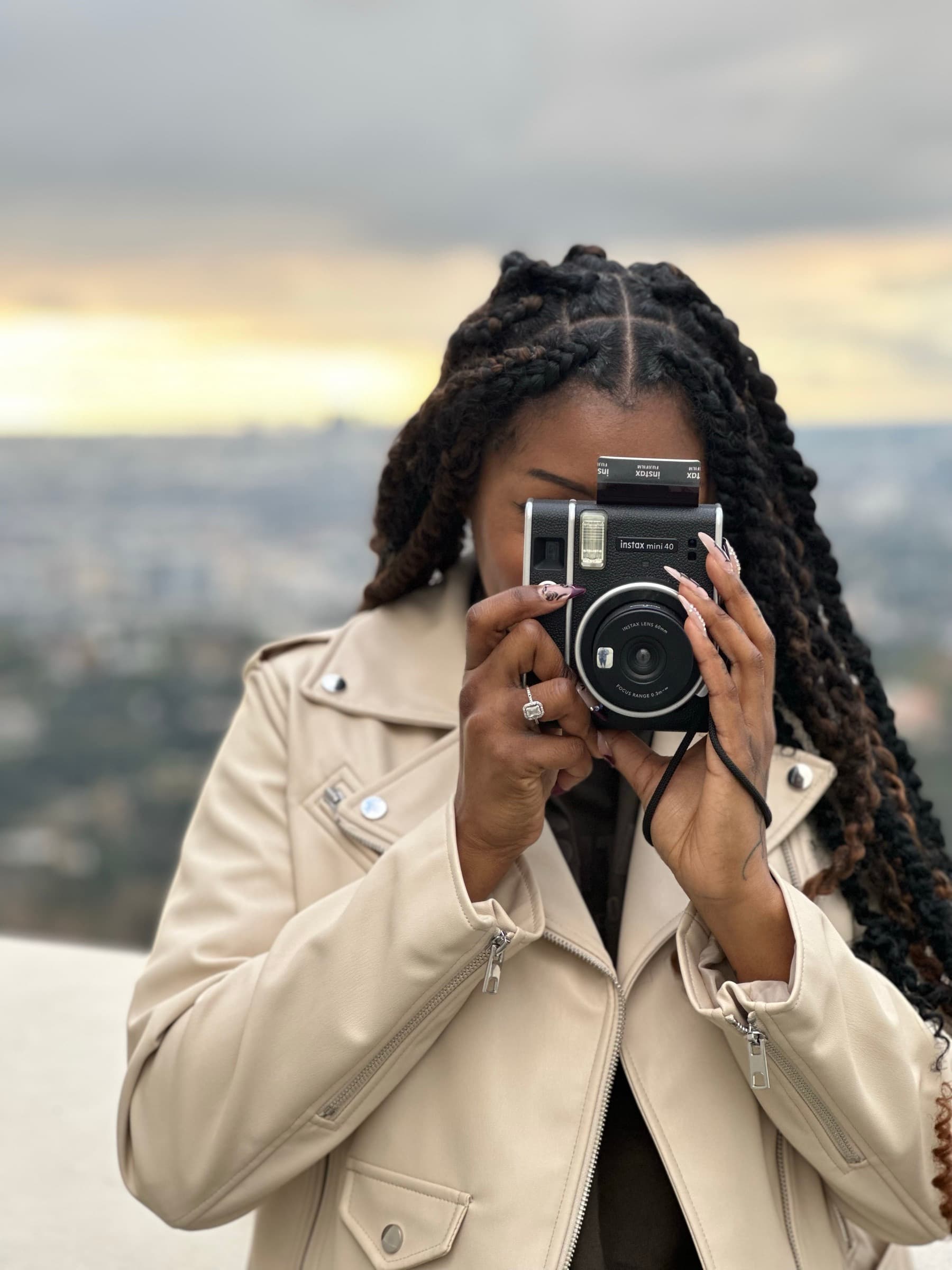 Woman raises an Instax instant camera to her eye, photographing the photographer, before a hazy city overlook